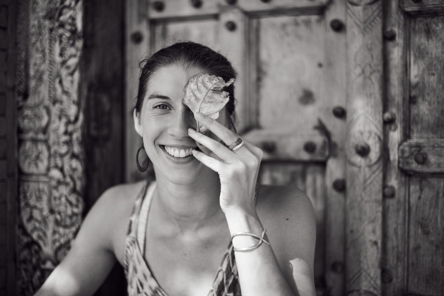 Smiling woman holding a leaf over one eye, wearing a bracelet, posed in front of a rustic wooden door.
Lotus Land.Photo