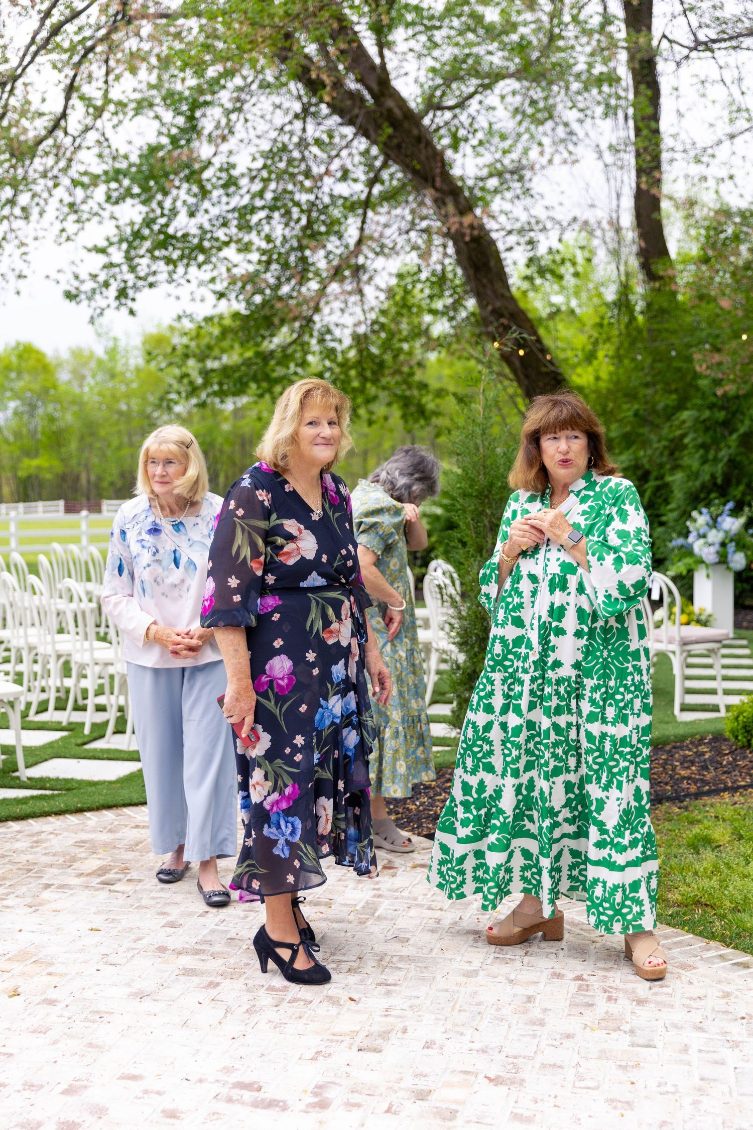 Wedding guests in colorful floral dresses walk through an outdoor garden ceremony venue with white chairs.