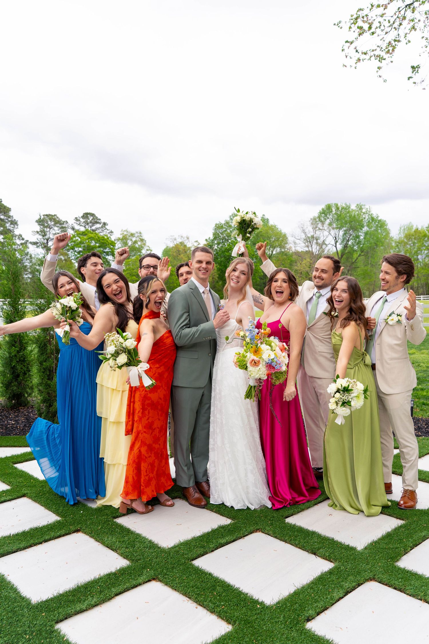 Joyful wedding party celebrating in colorful dresses and suits on geometric grass and stone patio.