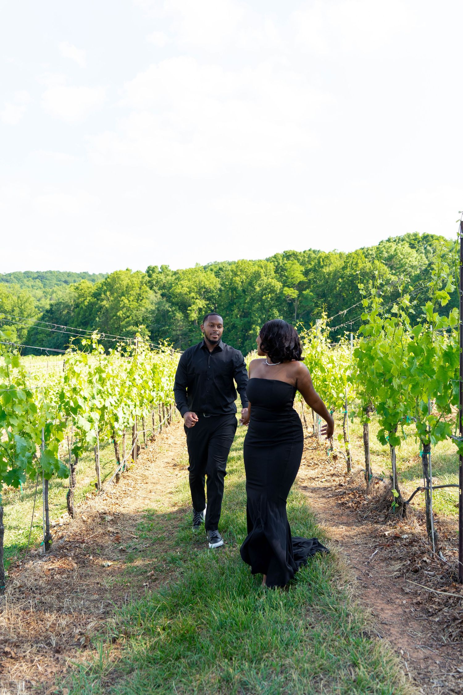 Couple in formal black attire walk together along a sunlit vineyard pathway.