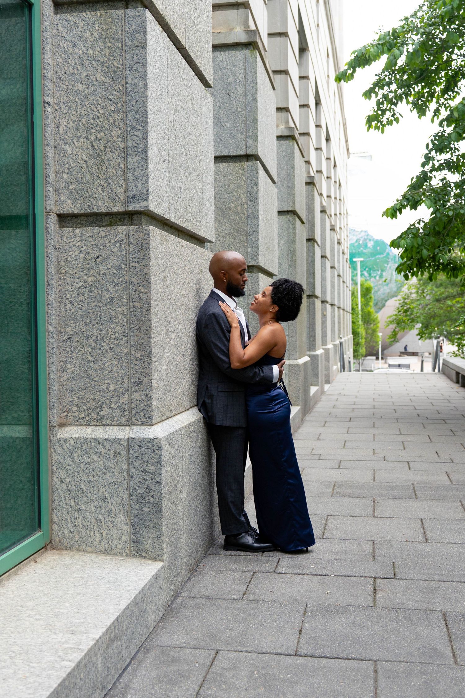 A couple in formal wear shares a romantic moment against a granite building wall on a city sidewalk.
