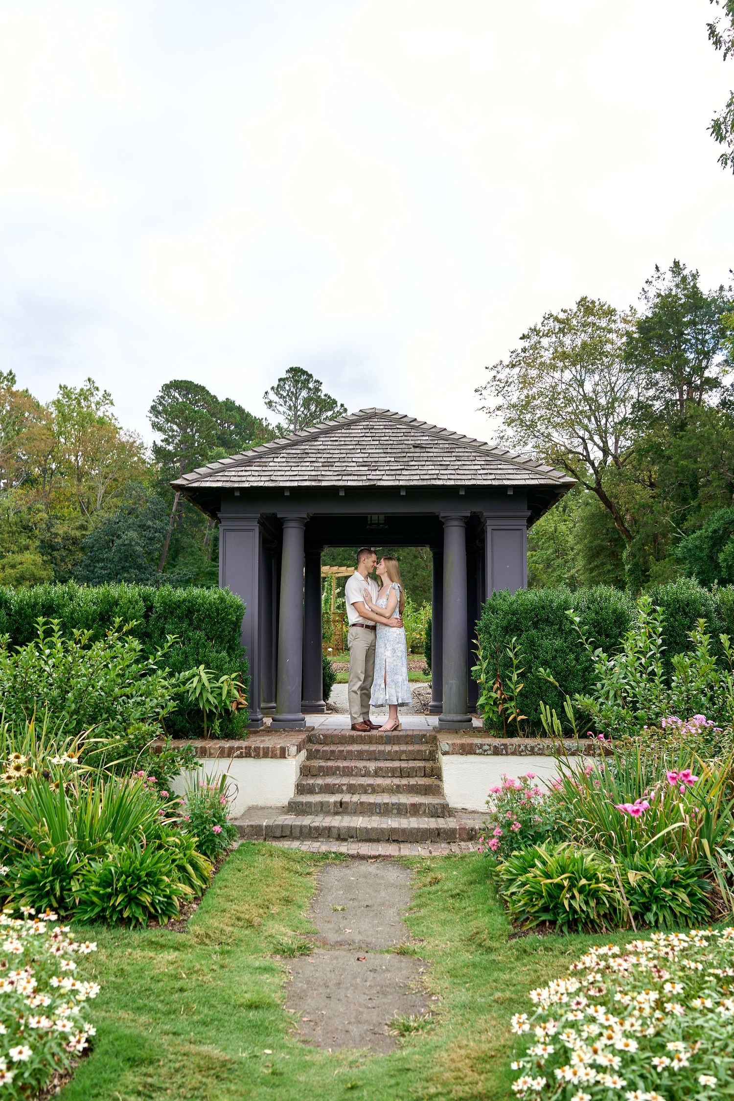 Romantic couple kissing under a classic dark pergola gazebo in a lush garden with pink flowers.