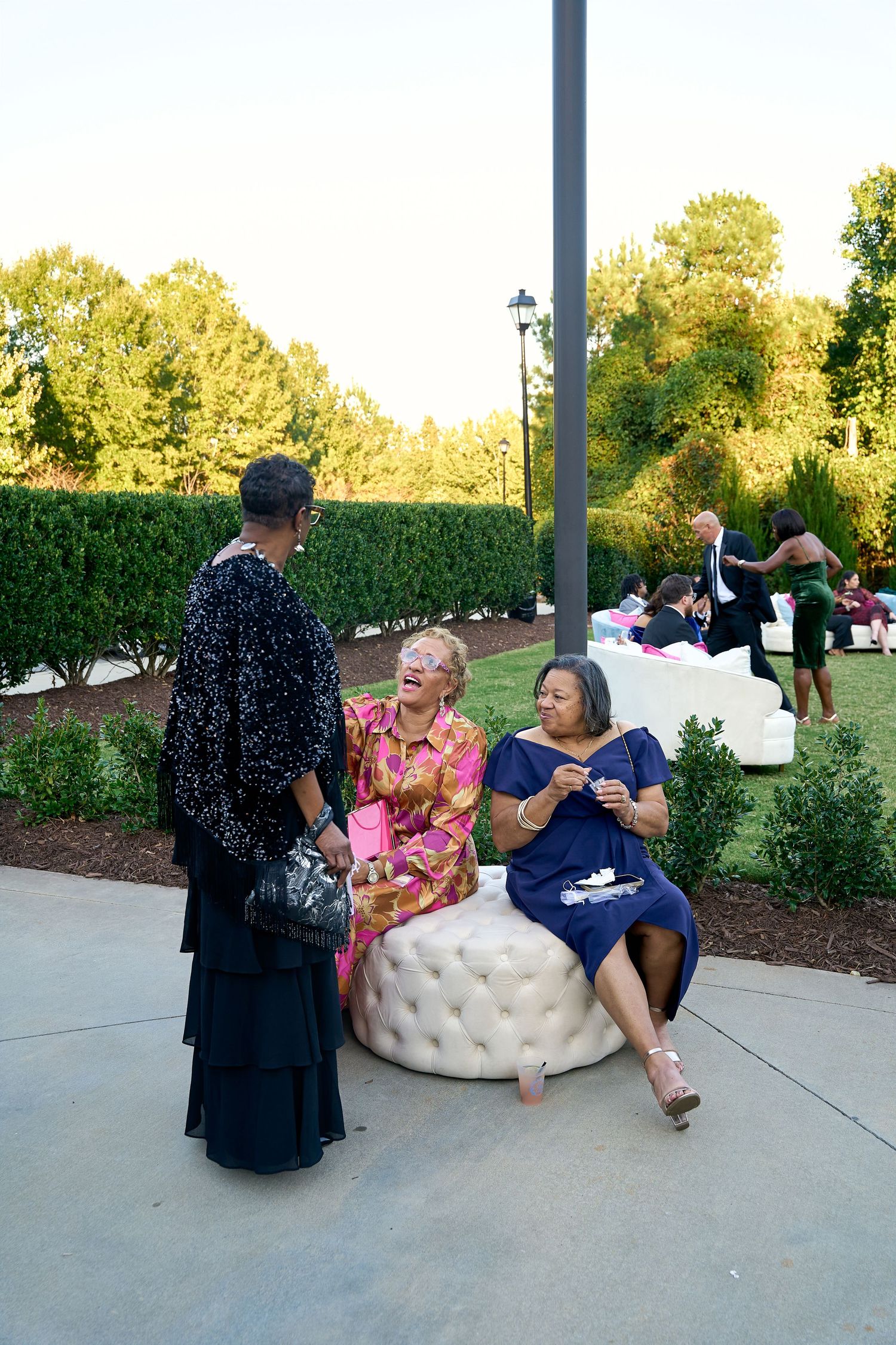 Three women in formal attire laugh and chat together at an outdoor wedding reception at dusk.