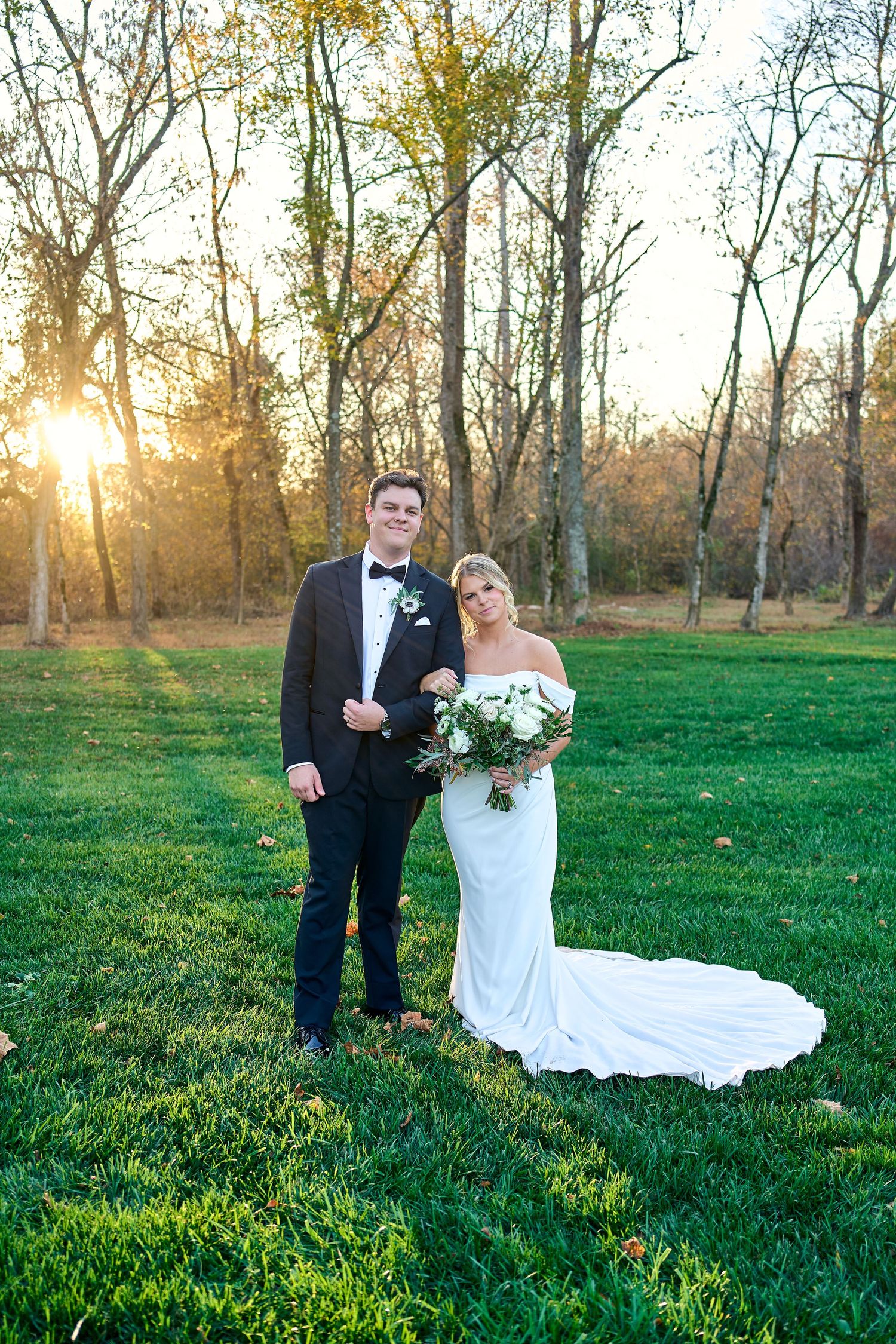 A couple in wedding attire stands on a lush green lawn at sunset with bare trees and golden sunlight in the background.