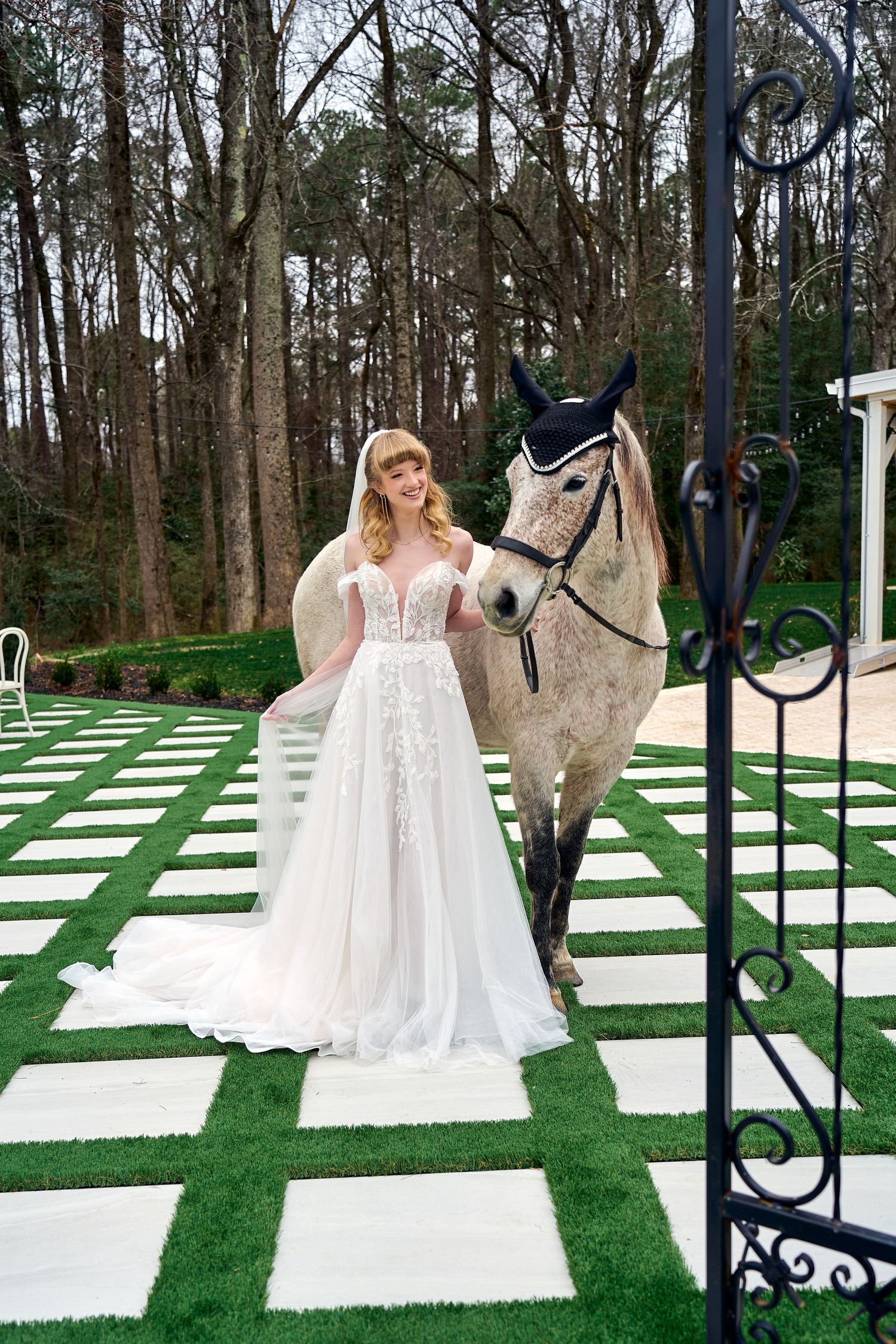 Wedding dress on display outdoors with a white horse on a decorative checkerboard lawn with trees in background.