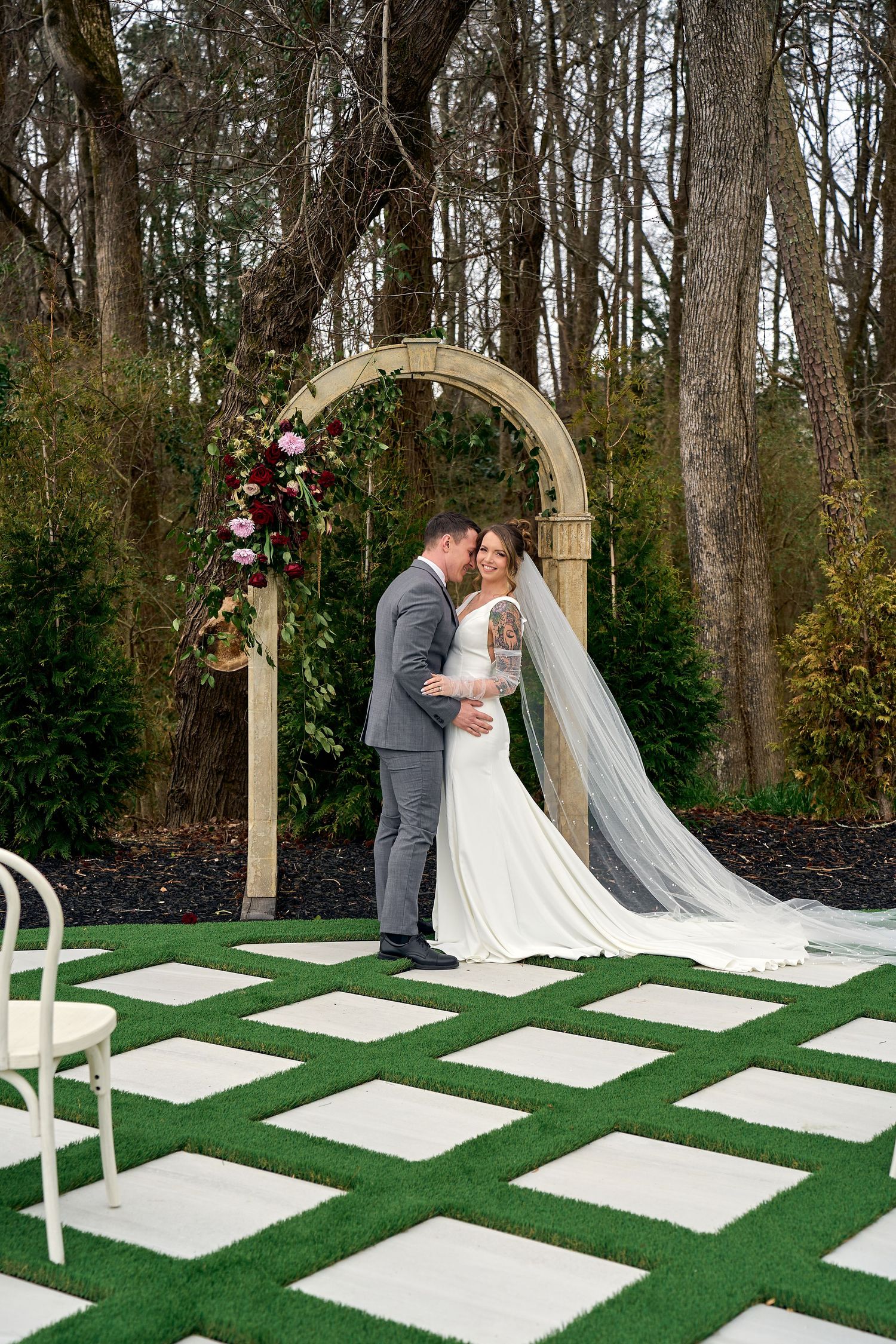 A romantic outdoor wedding ceremony setup features an arch decorated with pink flowers and white stone pavers on green grass.
