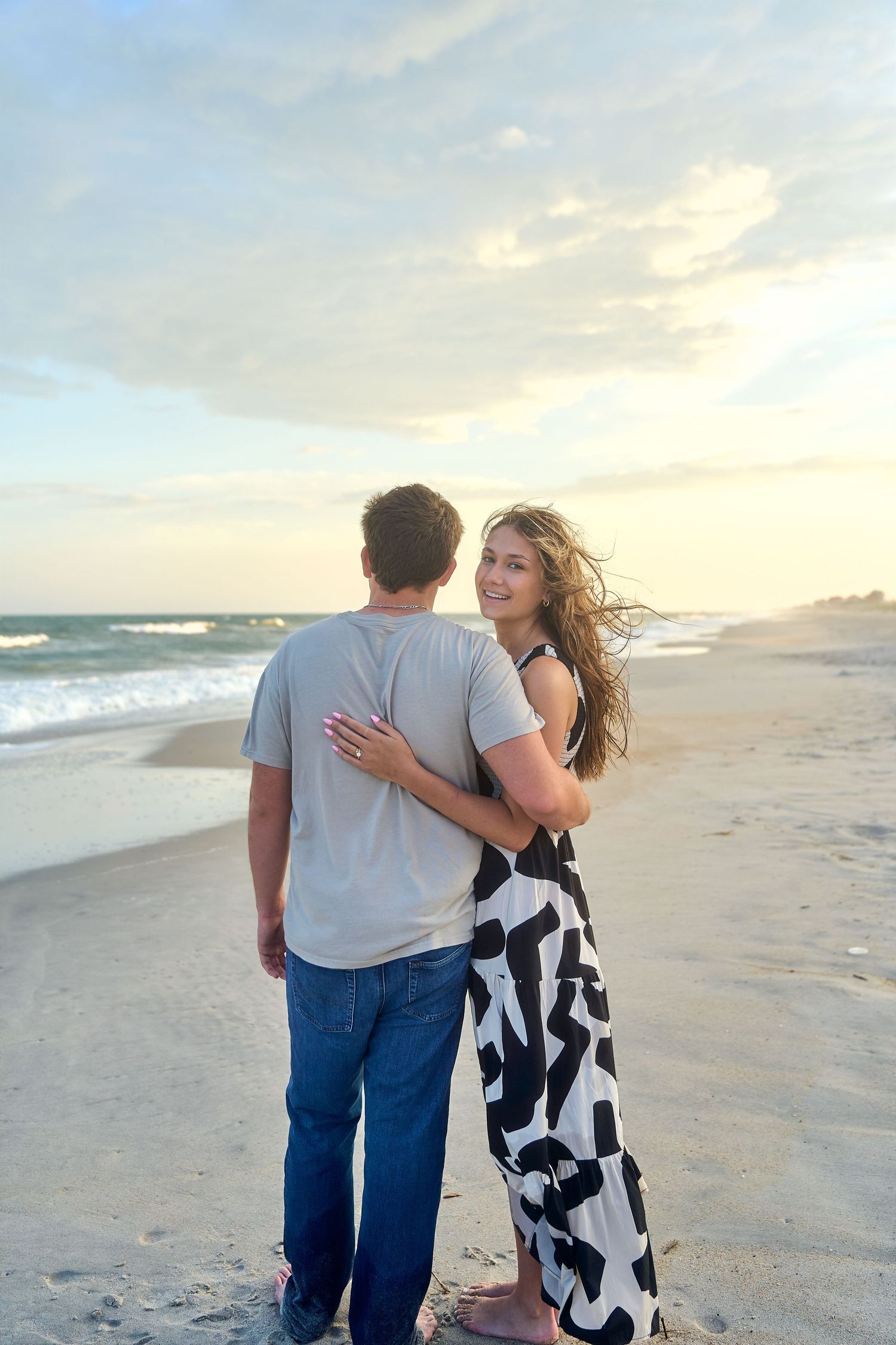 A couple embraces while walking on a sunny beach at sunset with waves crashing in the background.