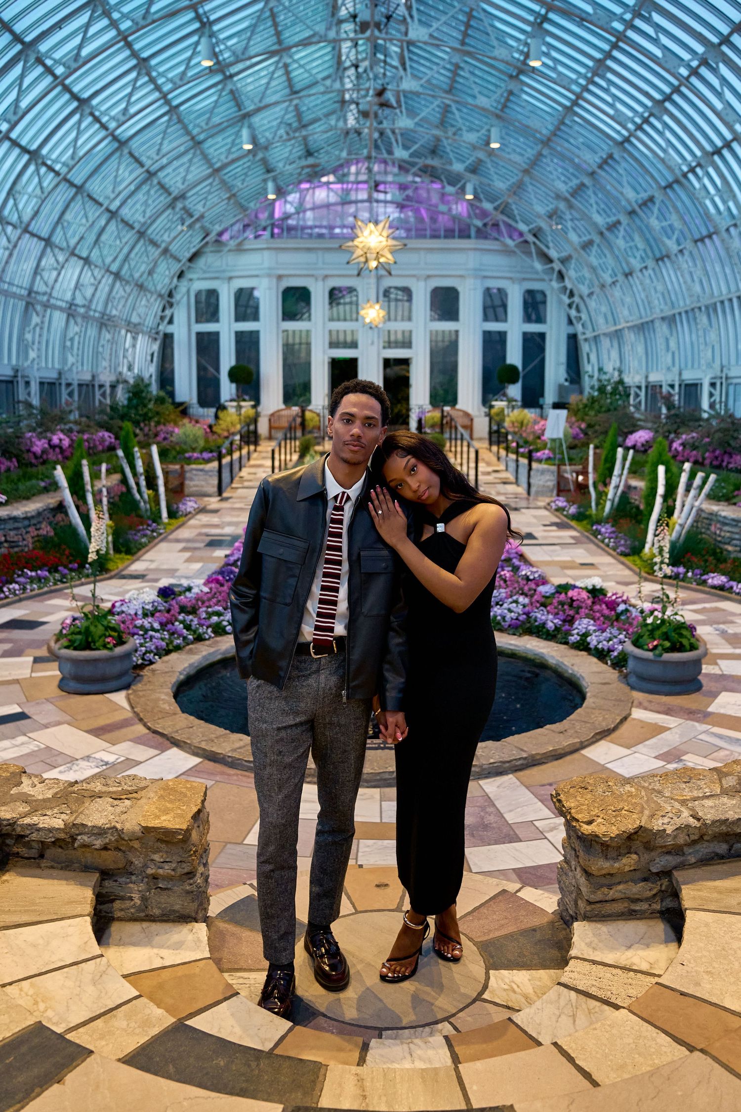 Couple posing together inside a grand glass conservatory with purple-lit dome and colorful flower beds.