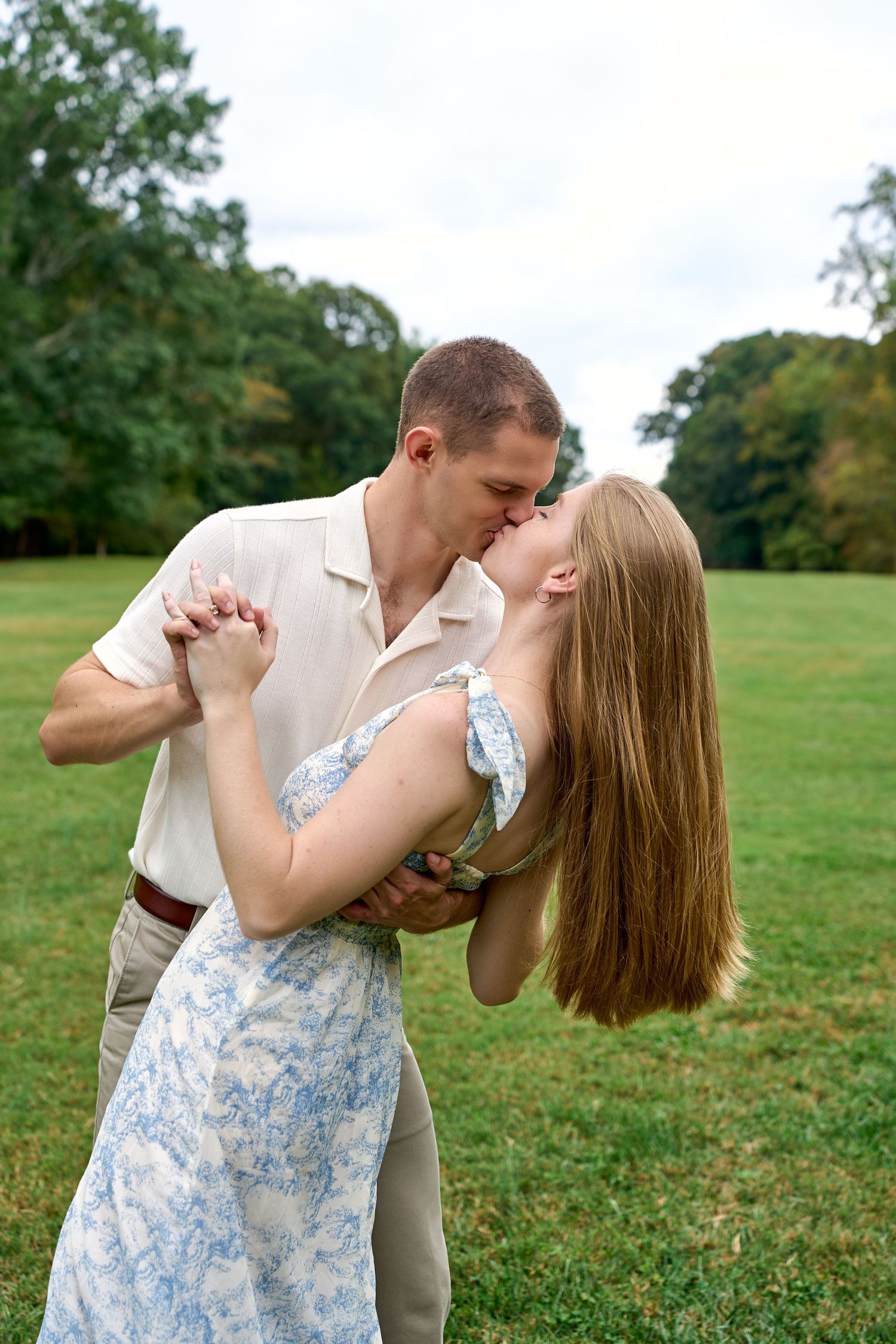 Couple sharing a romantic dip kiss on a green lawn with trees in background.