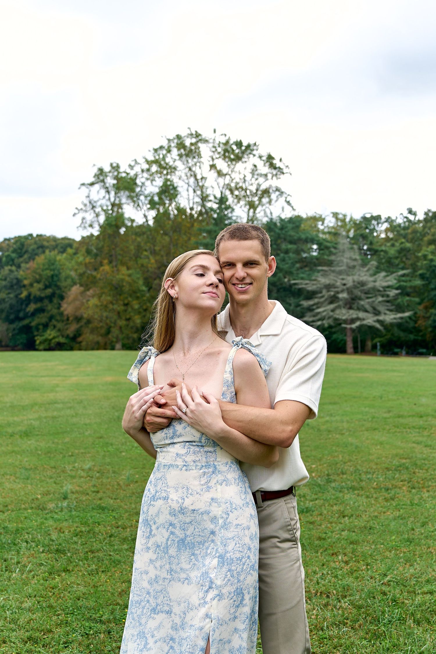 A couple embraces in a romantic moment on a vast green lawn with trees in the background. North Carolina Wedding Photographer.