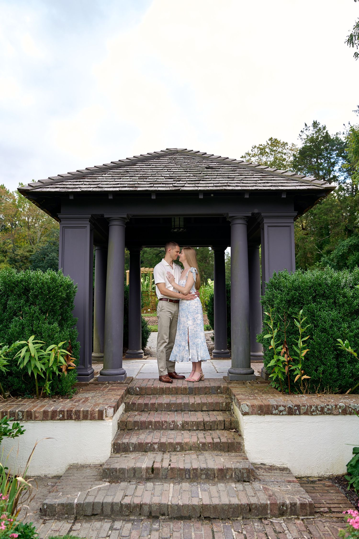 A couple shares an intimate moment under a gray pergola with stone steps and tropical plants. North Carolina Wedding Photographer.