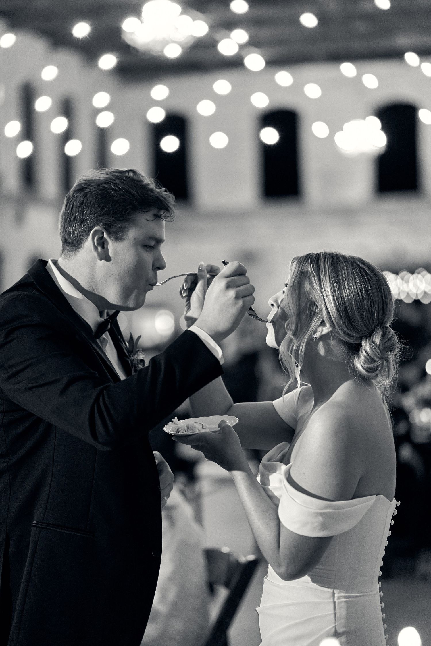 A wedding couple feeding each other cake during their reception with soft bokeh lights in the background.