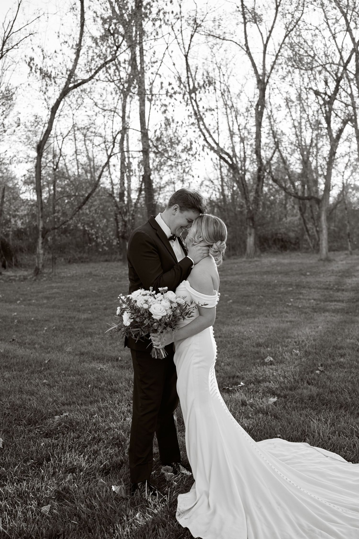 A tender black and white wedding photo shows a romantic moment between newlyweds embracing in a wooded outdoor setting.