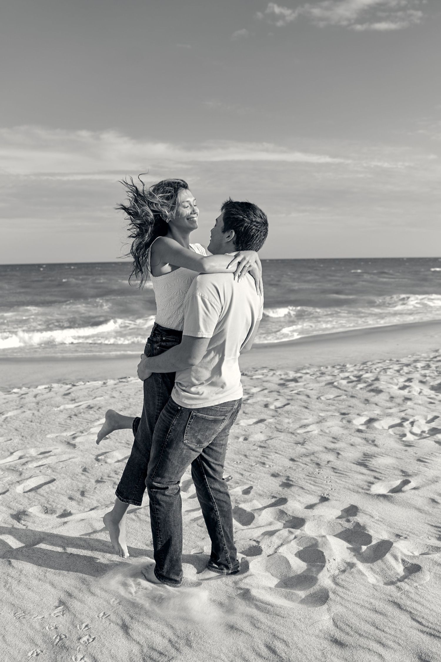 A couple embraces on a sandy beach with ocean waves and cloudy sky in black and white.