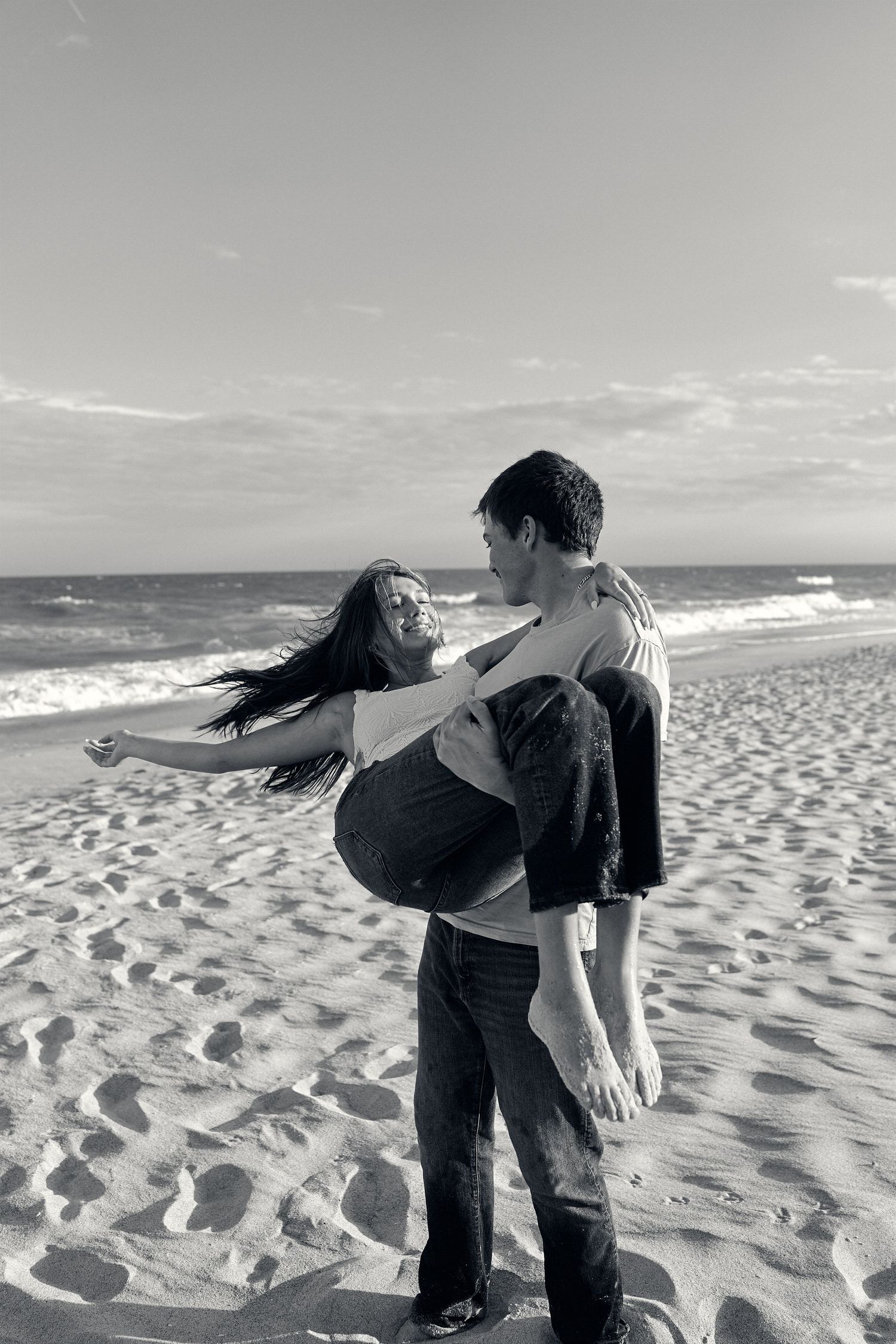 A romantic black and white moment between two people dancing together on a sandy beach with ocean waves in background.