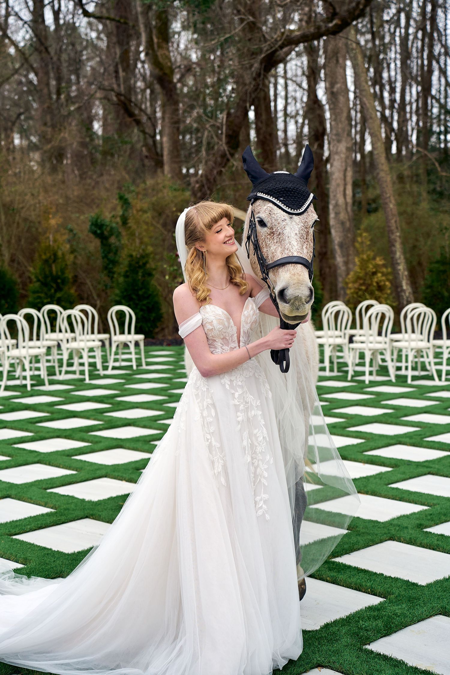 A white horse and someone in a flowing lace wedding dress pose in front of white garden chairs on a checkered lawn.