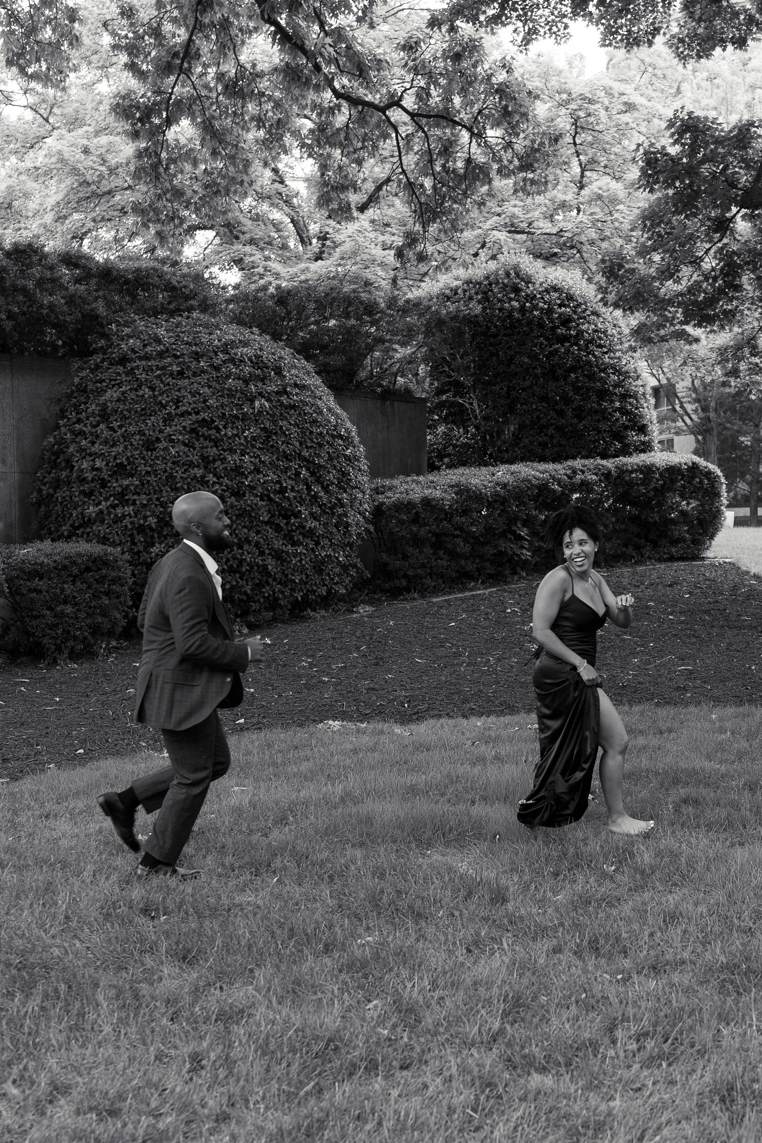 A couple in formal wear playfully runs across a manicured lawn in a black and white photo.