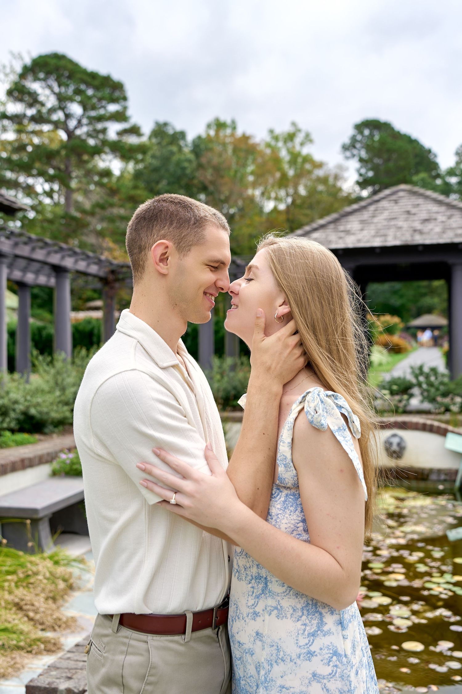 Smiling couple leaning in for a kiss near a tranquil garden pond with lily pads and pergola.