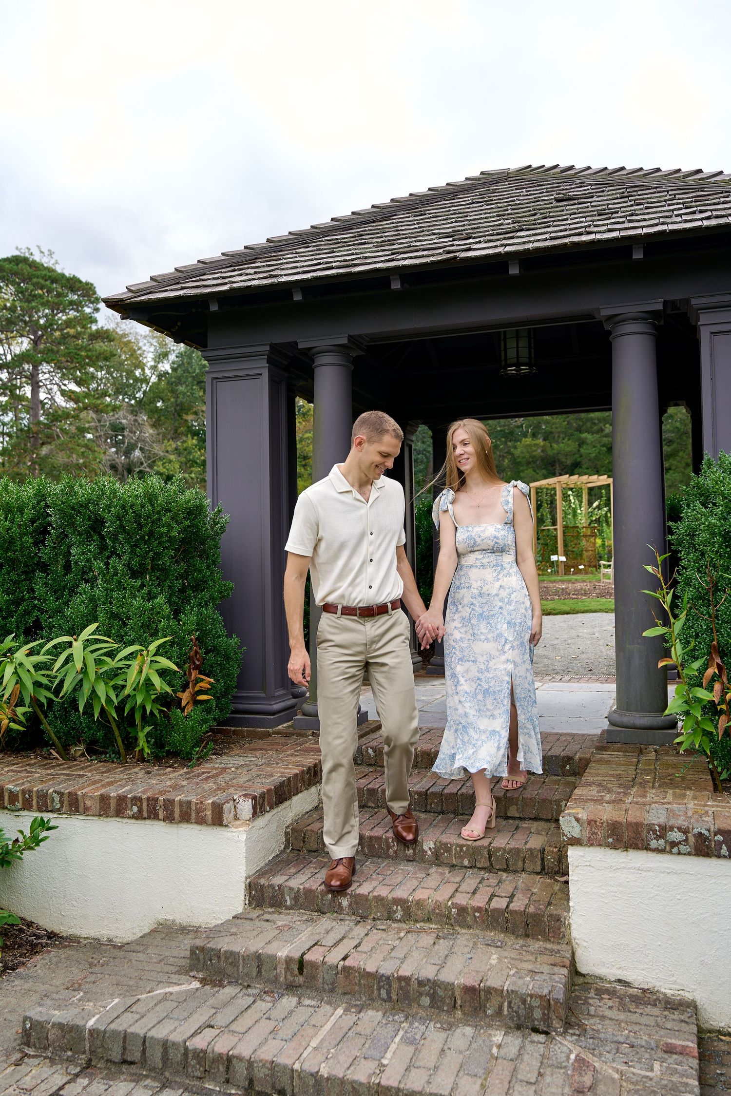 Couple holding hands walking down brick steps from a garden gazebo with columns and lush greenery.
