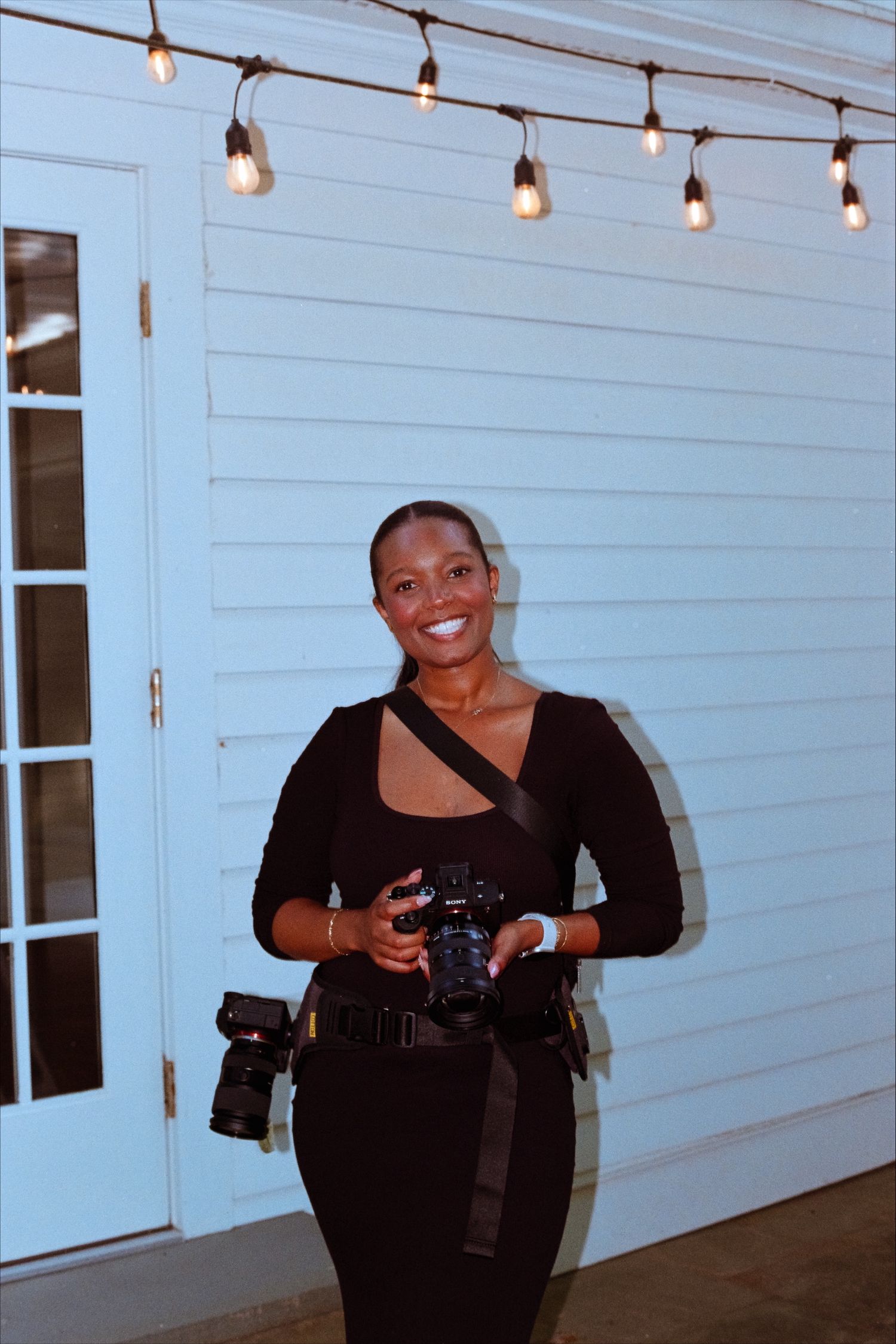 Smiling female wedding photographer in black outfit holding cameras under string lights at night.