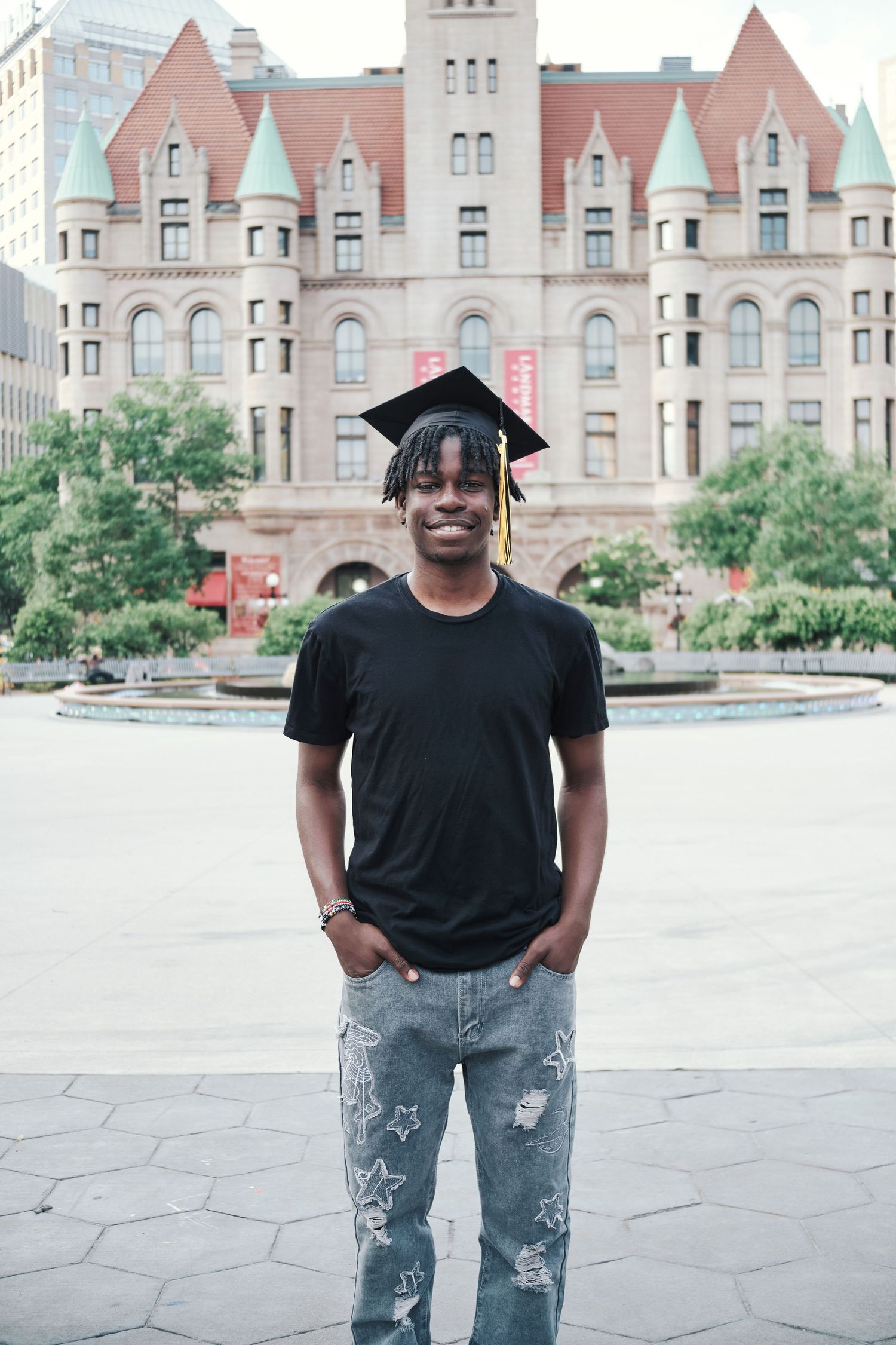 Young man in graduation cap and jeans smiling in front of historic university building.