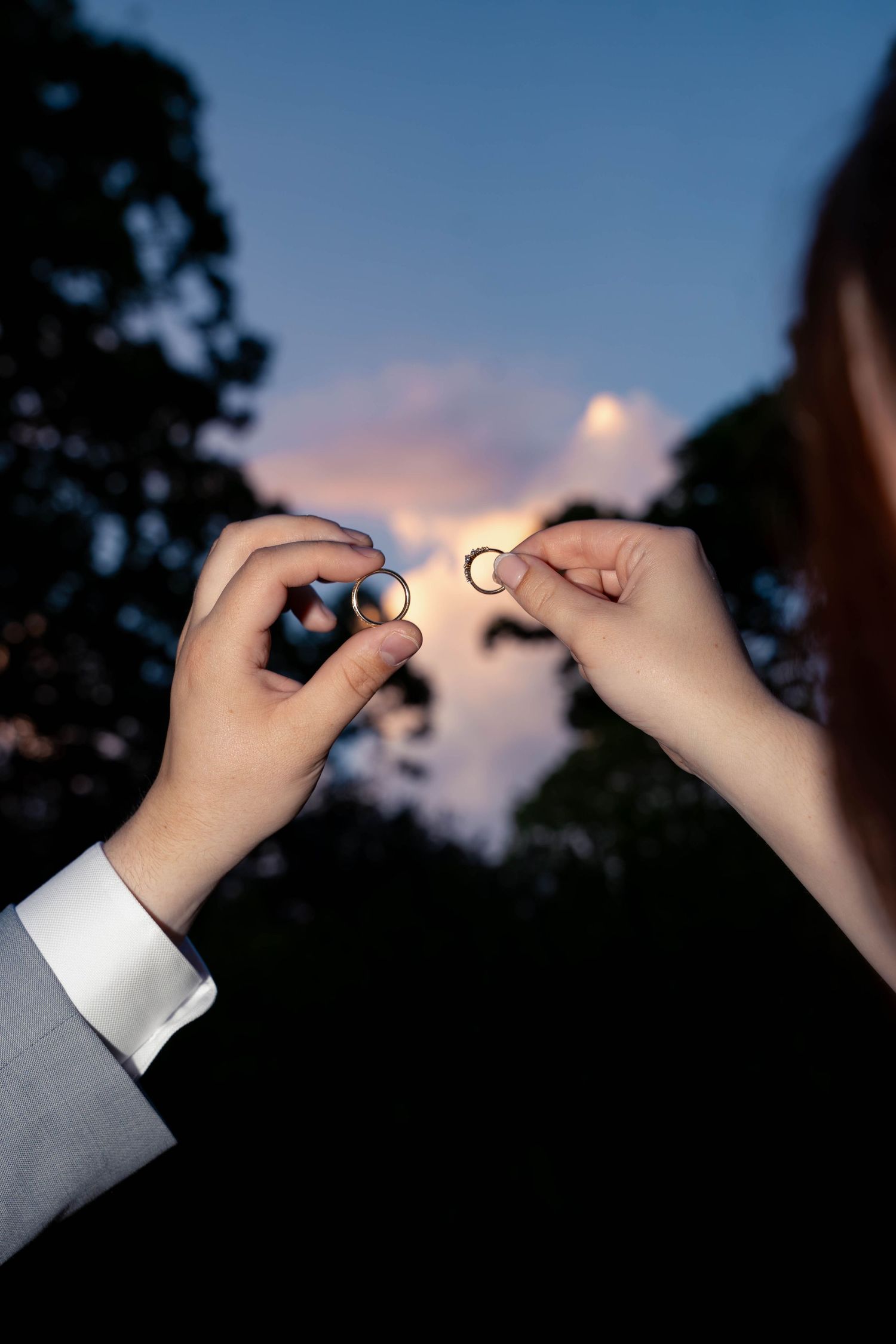 Bride and groom holding wedding rings up against twilight sky at dusk.