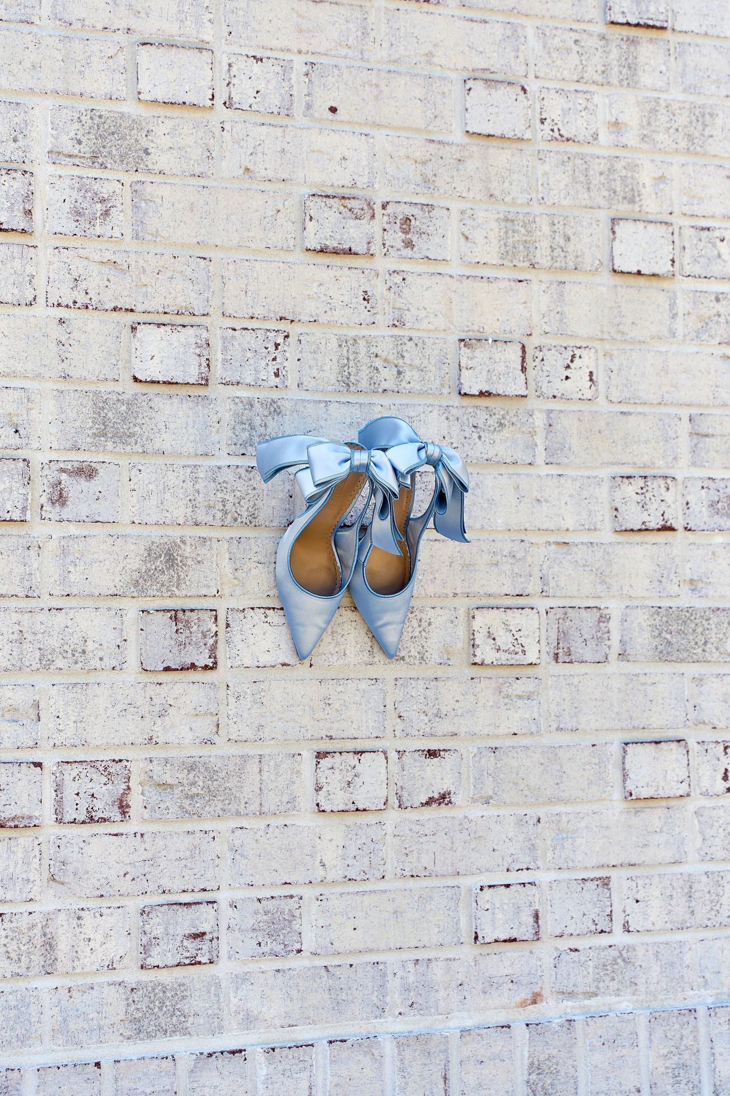 Blue satin bow heels hanging on rustic brick wall as wedding detail shot.