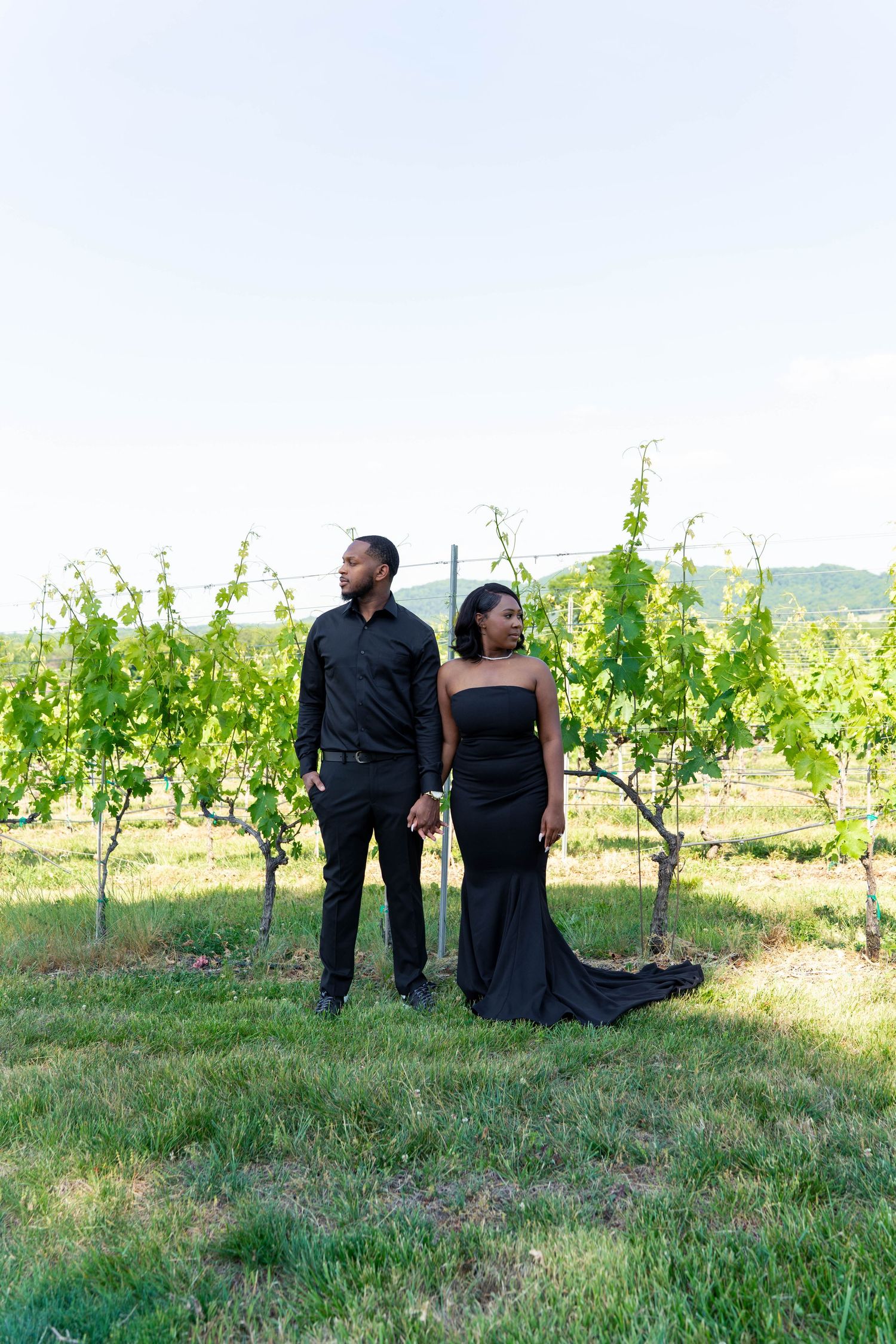 Elegant couple in all-black attire holding hands and posing among green vineyard rows under bright sky.