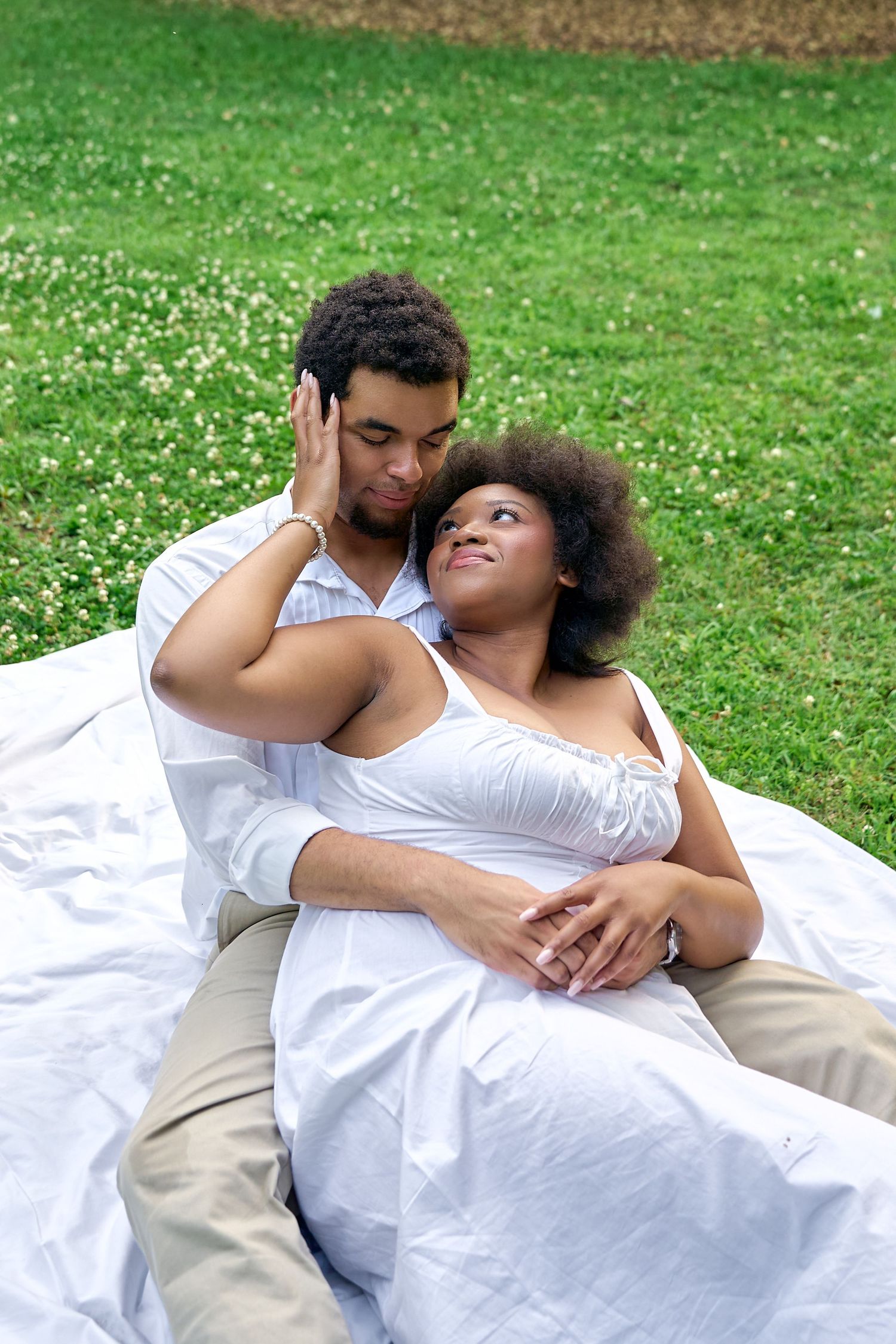 Couple relaxes on white blanket in grassy field, sharing tender moments together.