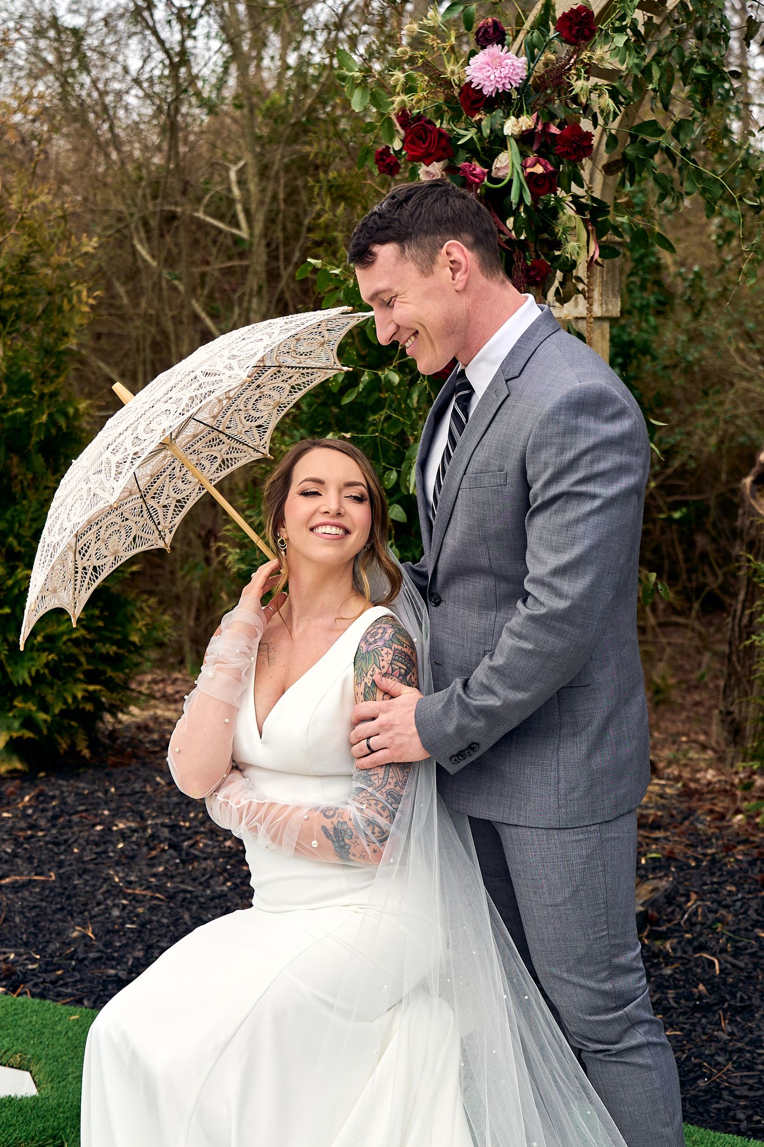 A couple shares a joyful moment at their outdoor wedding ceremony with a vintage lace umbrella and flowing white dress.