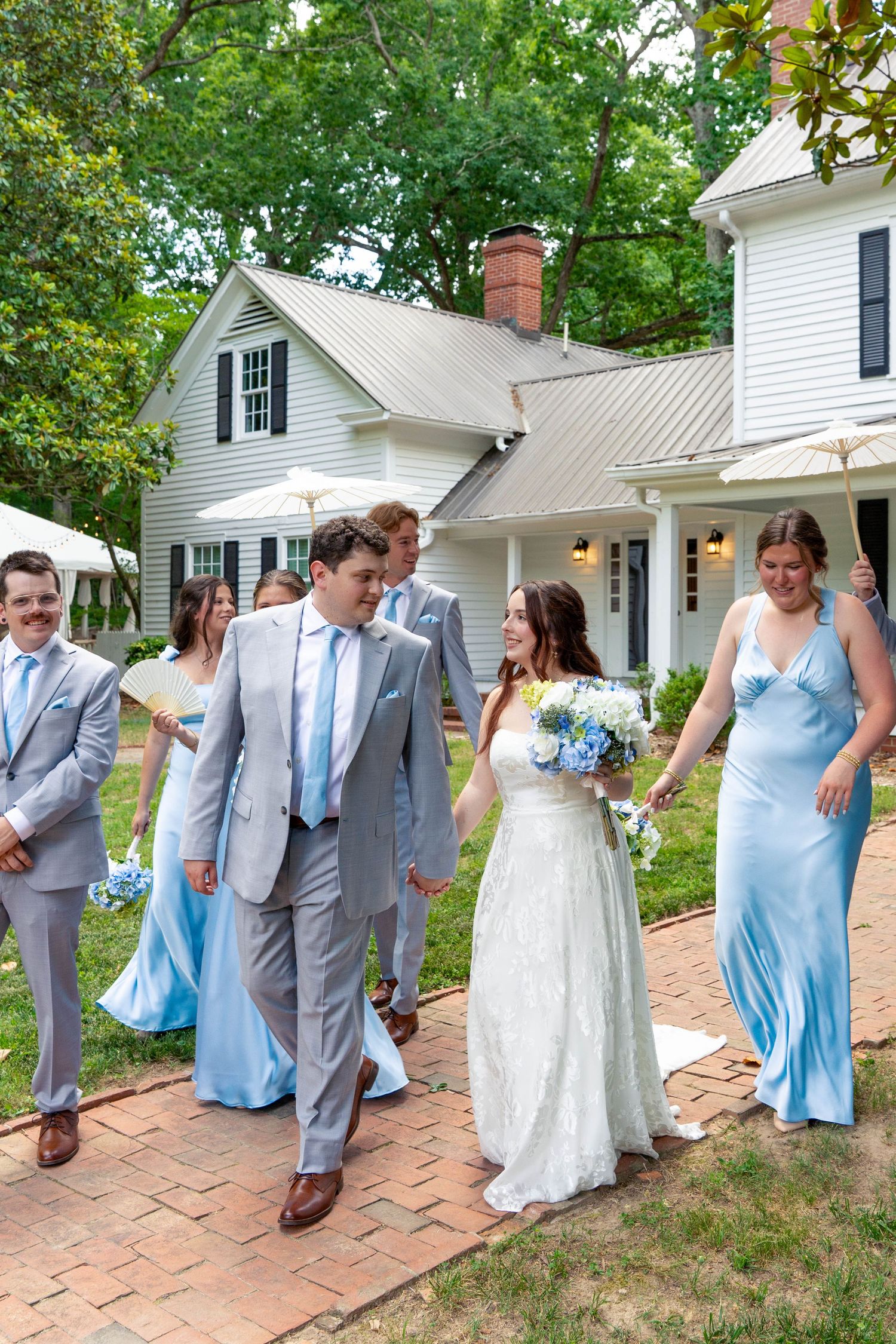 Bride and groom lead wedding party in blue and gray attire along brick path at white farmhouse.