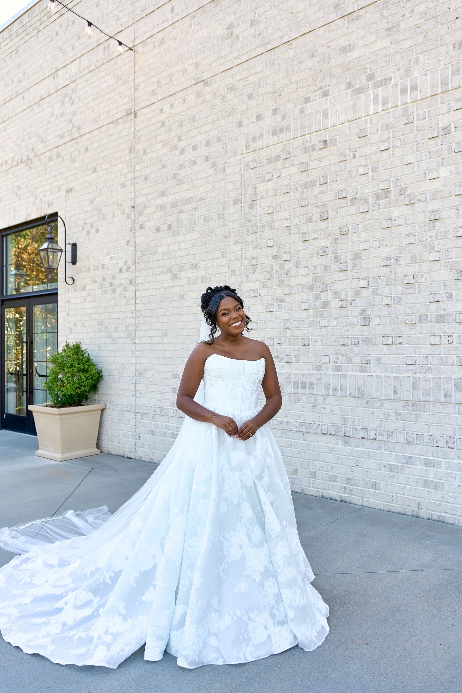 Bride in strapless lace ballgown wedding dress with long train poses outdoors against white brick wall.