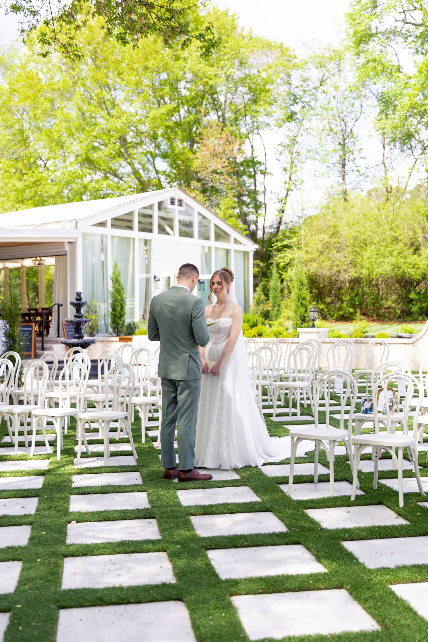 Bride and groom standing together at outdoor wedding venue with white chairs and greenhouse.