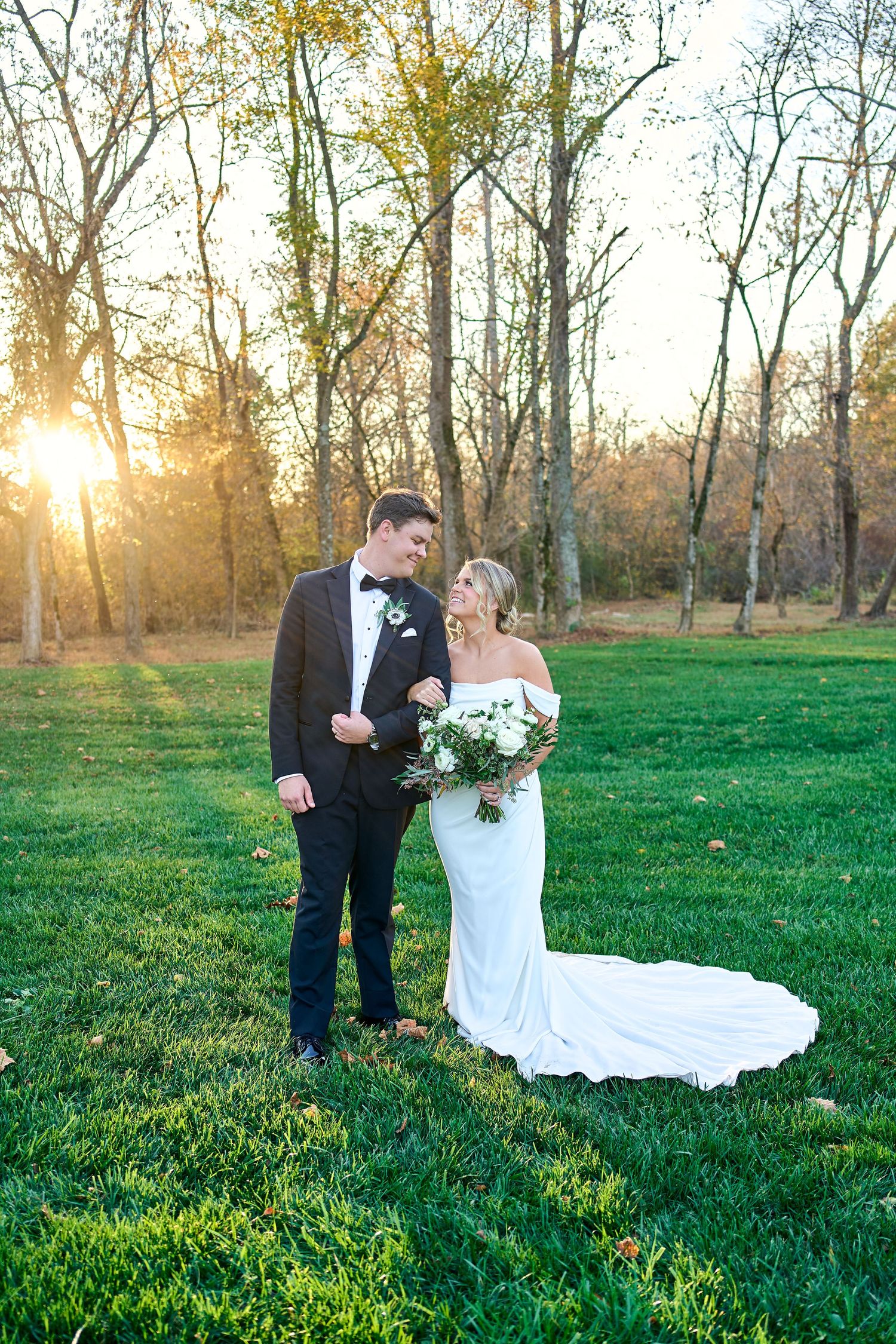 A couple in formalwear share a romantic moment in a sunlit field with autumn trees during sunset golden hour.