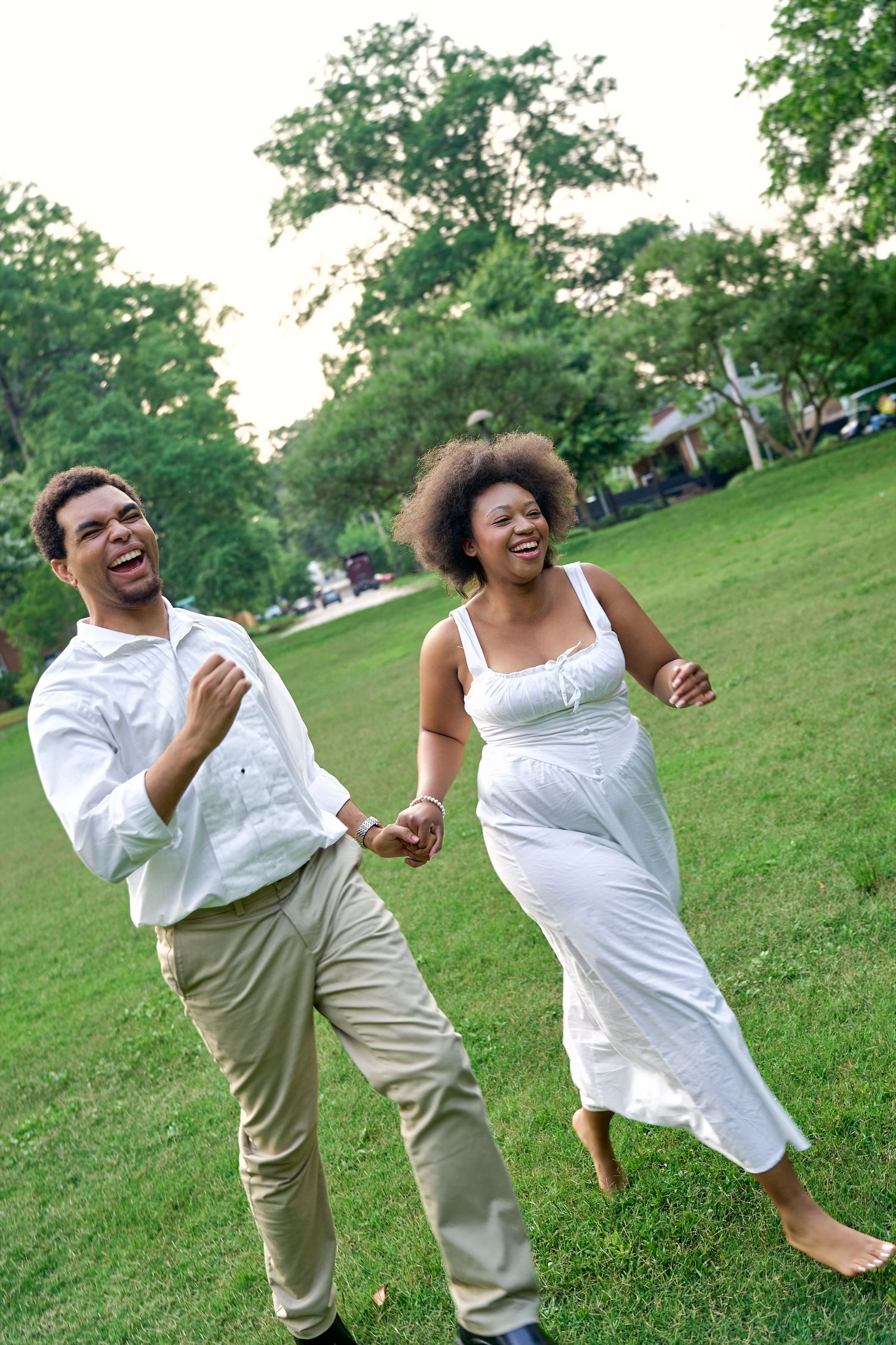 Two people in white and khaki outfits dancing joyfully on a grassy hillside on a sunny day.