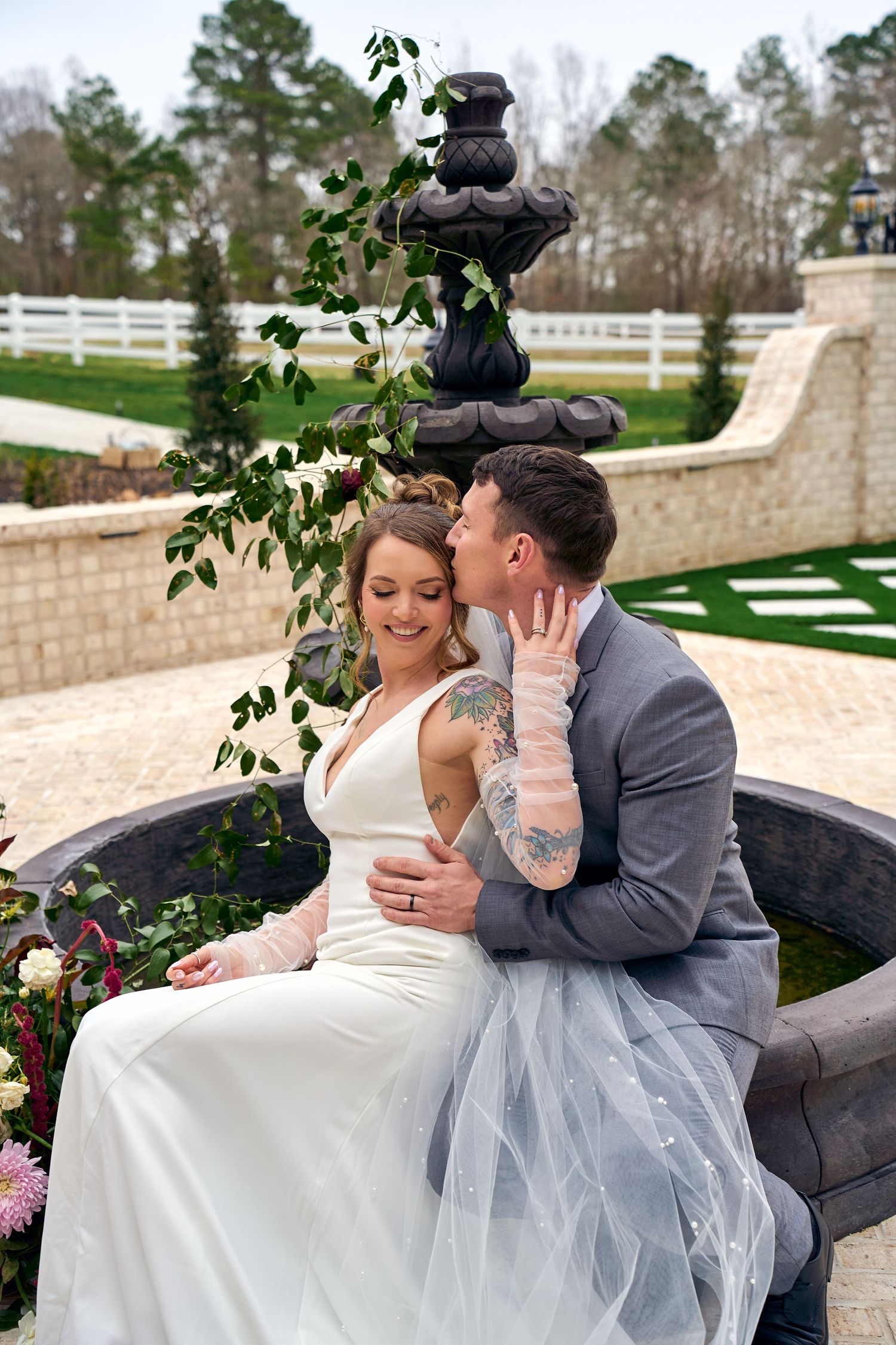 Couple in wedding attire embrace on stone fountain ledge surrounded by landscaped garden and white fence backdrop.