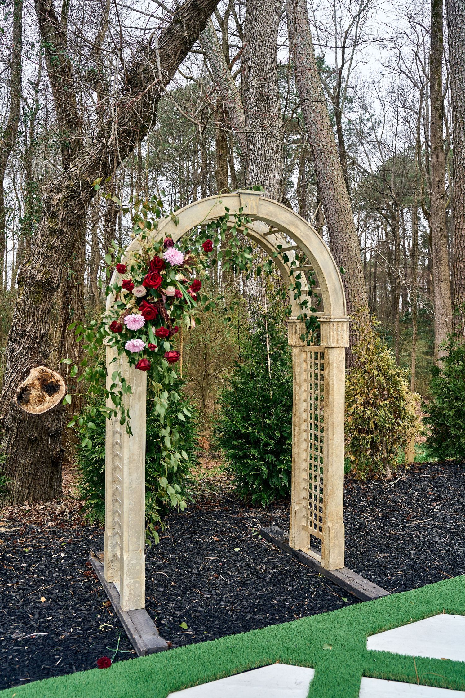 A wooden garden arch decorated with red and white roses and greenery stands in a wooded area with black mulch and turf.