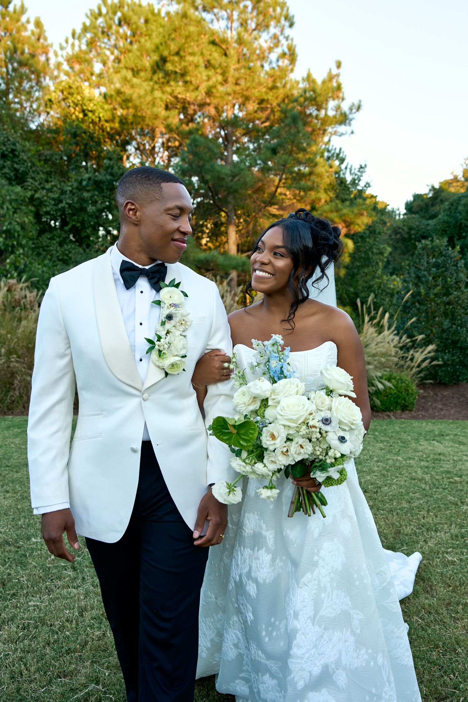 Smiling bride holding white and blue bouquet walks with groom in white tuxedo in garden setting.