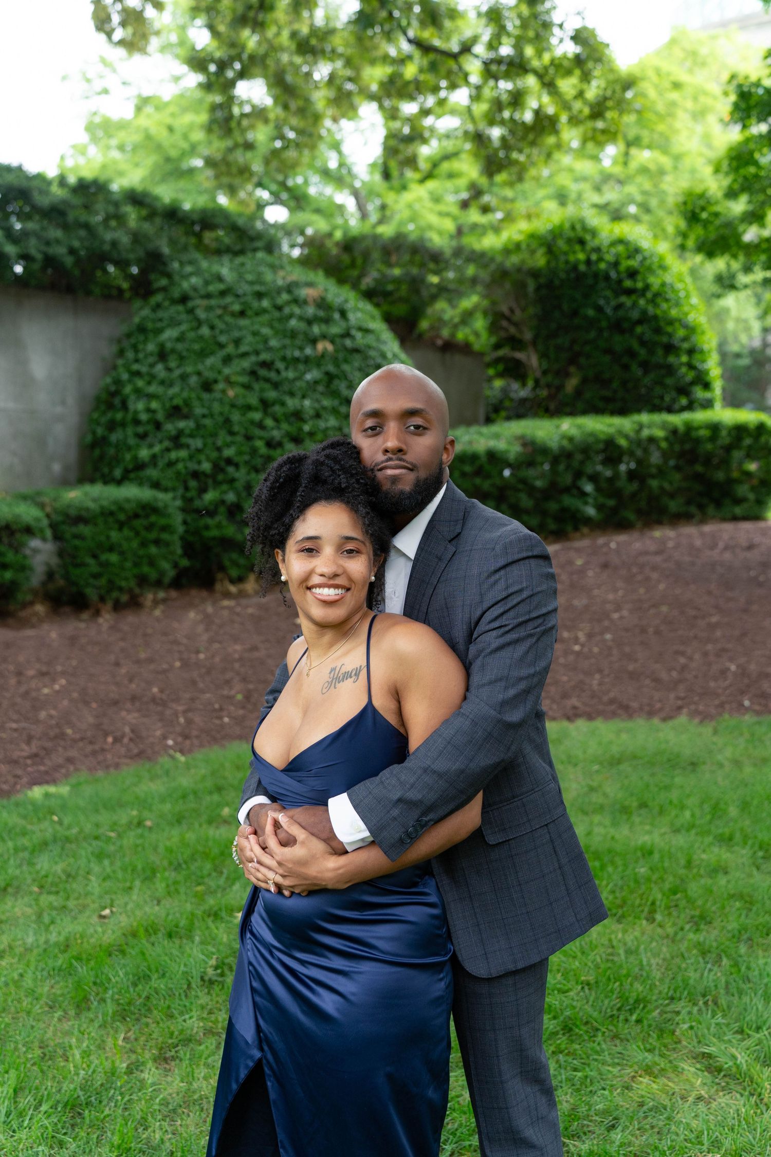 A smiling woman in a navy gown is embraced from behind by a man in a gray suit in a garden.