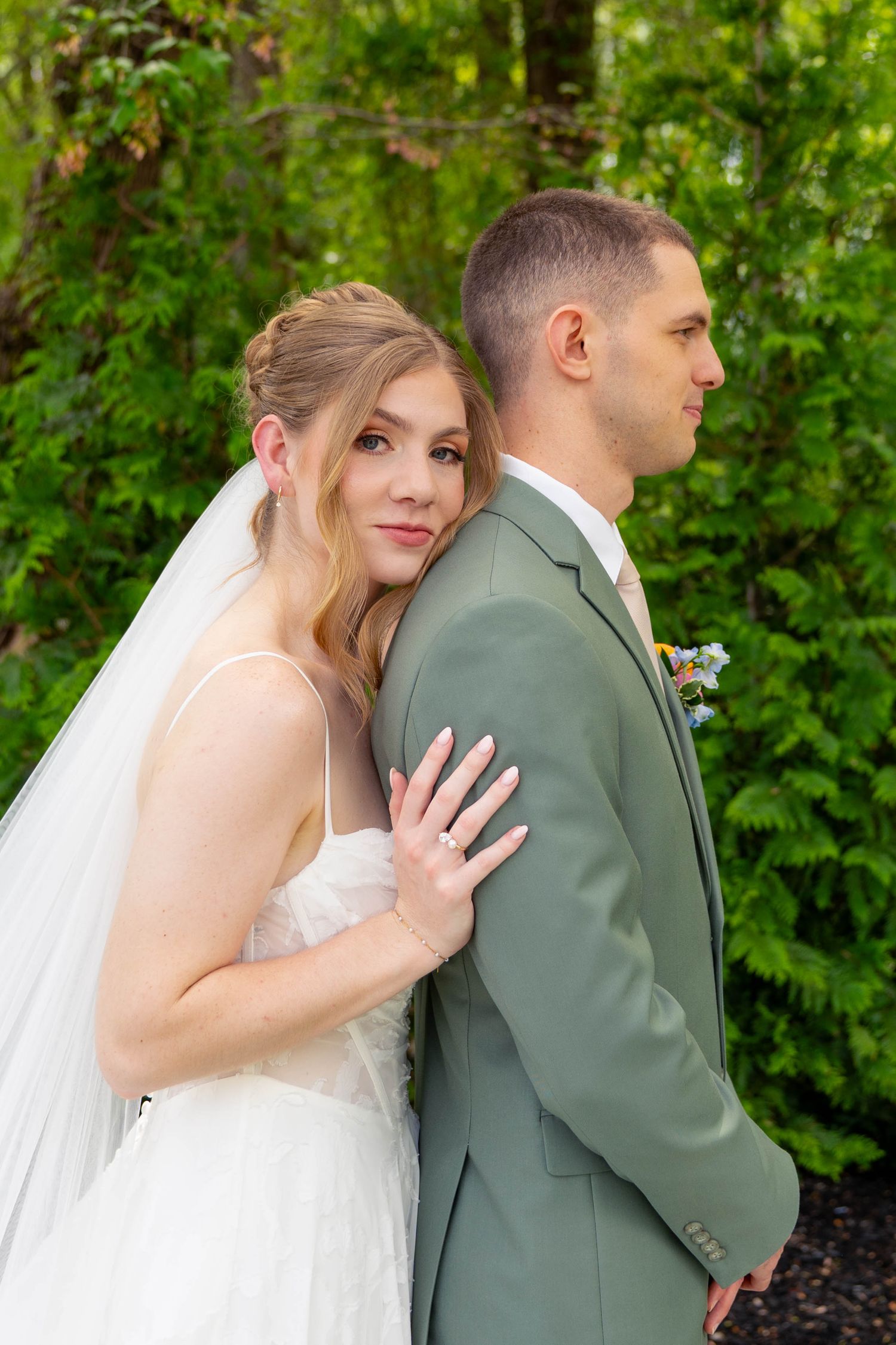 Bride in white gown and veil leans against grooms sage green suit jacket in a lush garden setting.