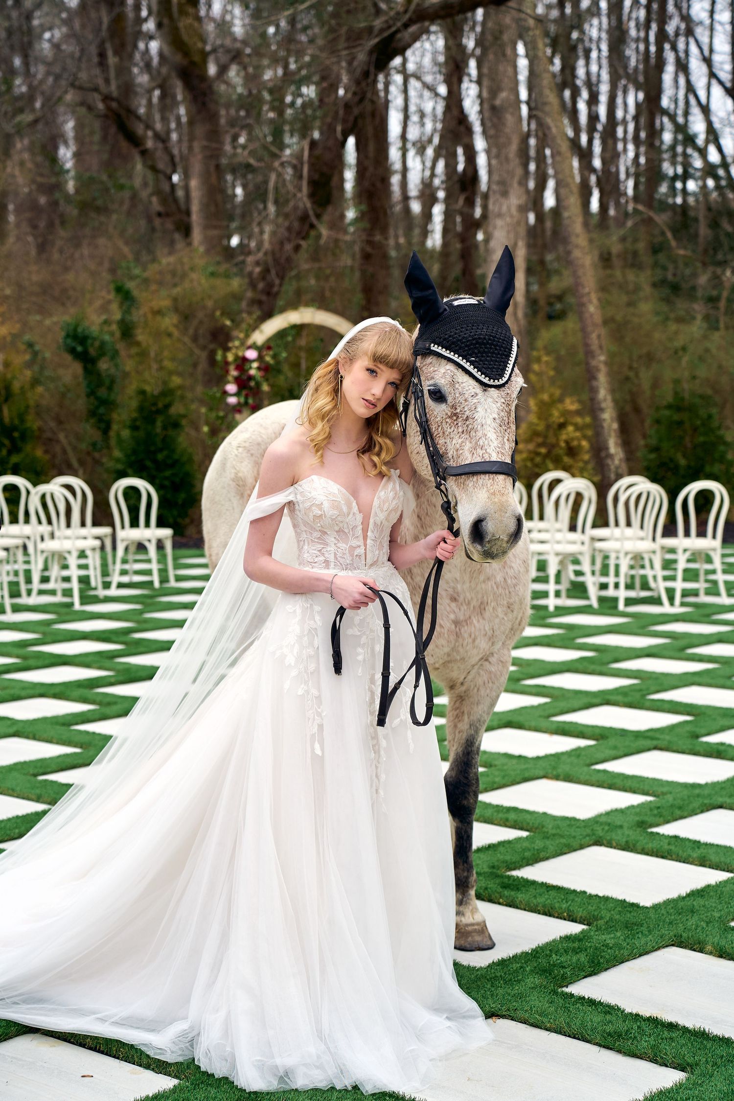 Elegant bride in a flowing white gown poses with a grey horse on a checkerboard lawn with white chairs at an outdoor venue.