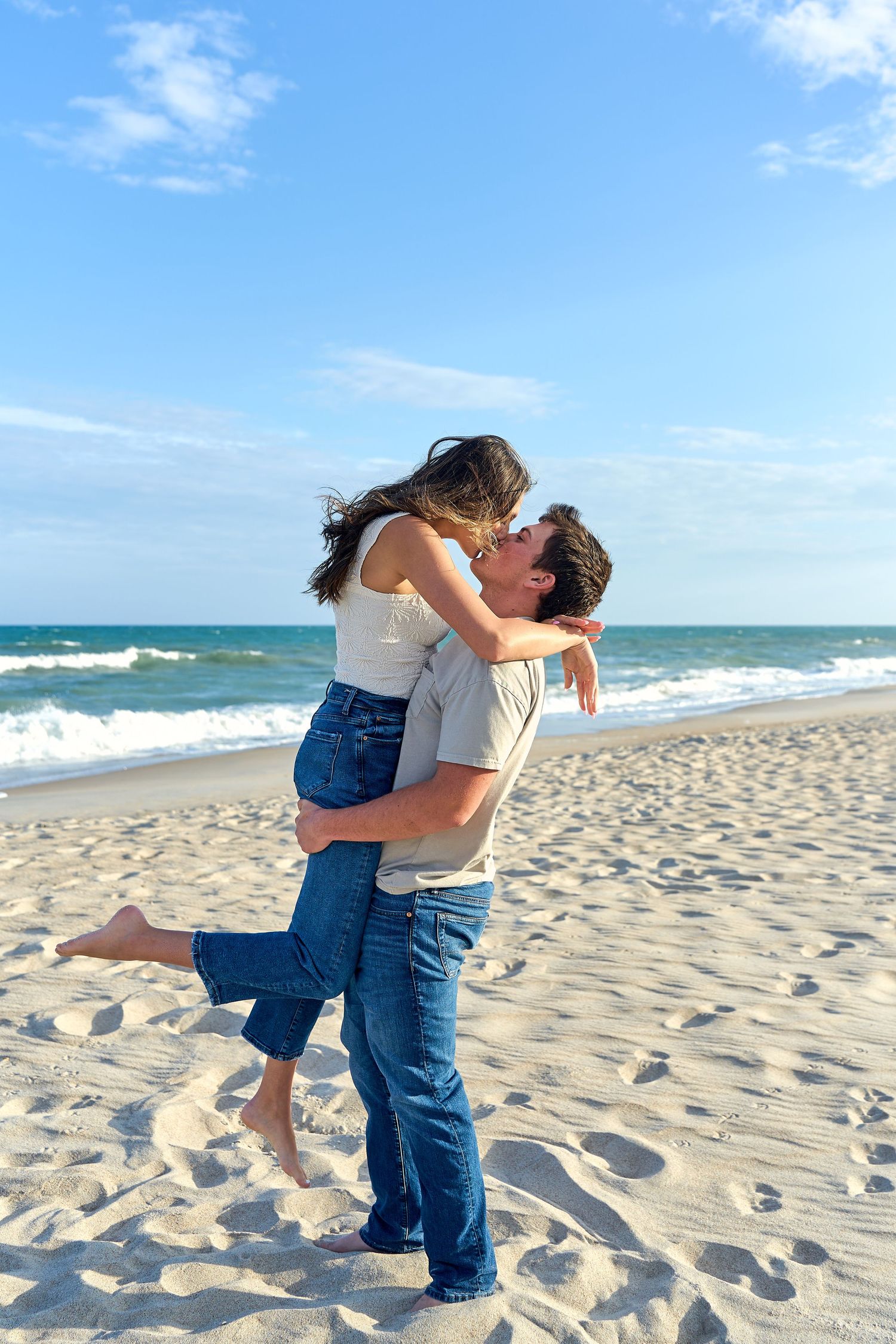 A romantic couple embracing on a sunny beach with ocean waves and blue sky in the background.