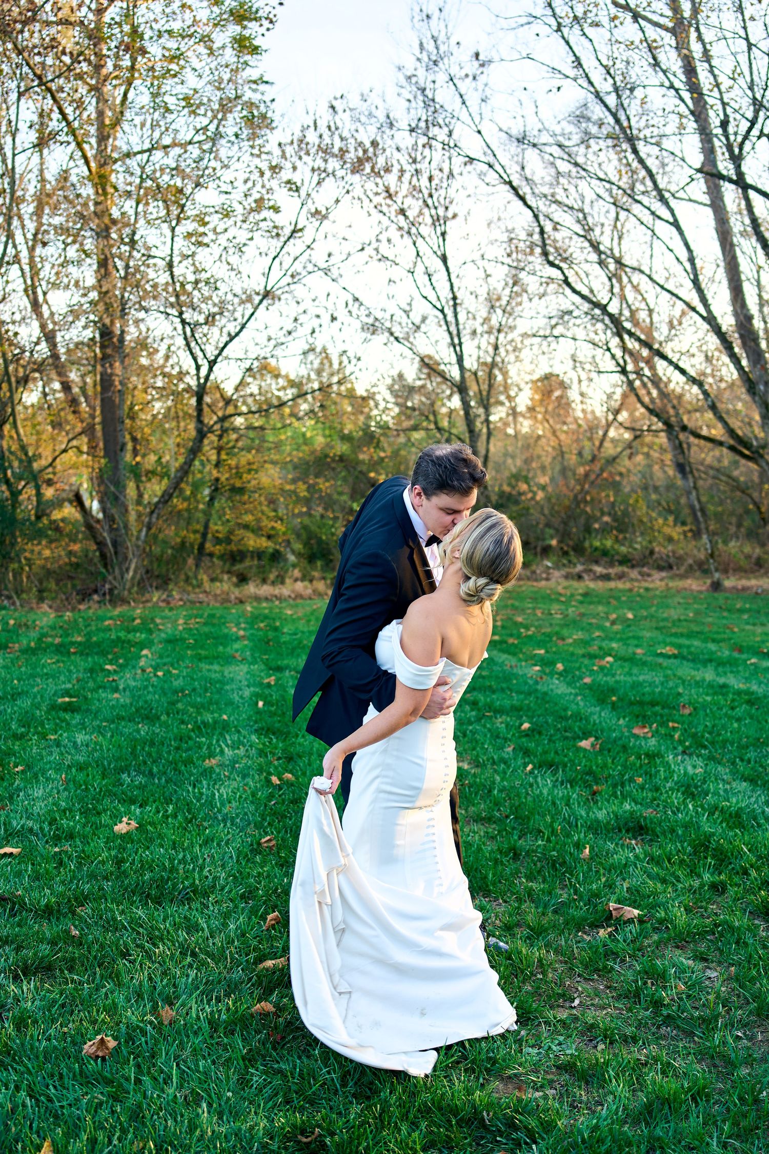 A couple shares a romantic dip kiss on a green lawn surrounded by autumn trees at sunset.