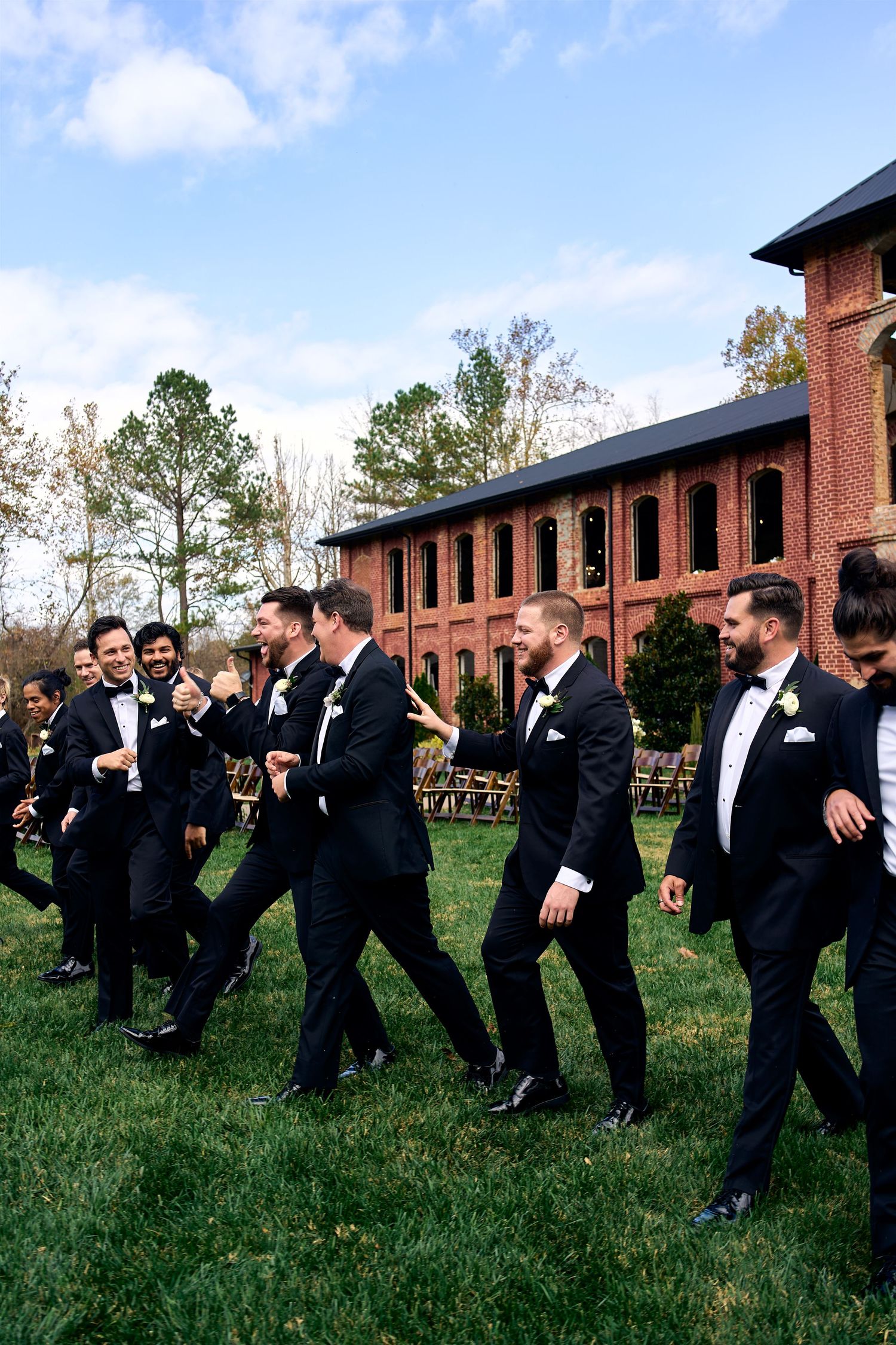 A group of groomsmen in black tuxedos walk and laugh together on grass outside a brick building on a sunny day.