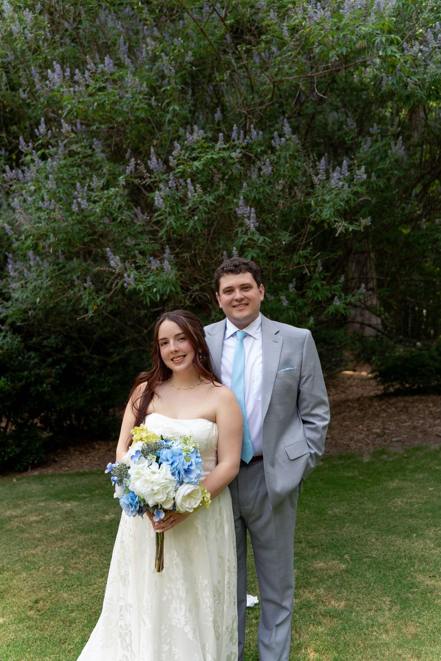Bride and groom posing together in a lush garden, she holds a blue and white bouquet.