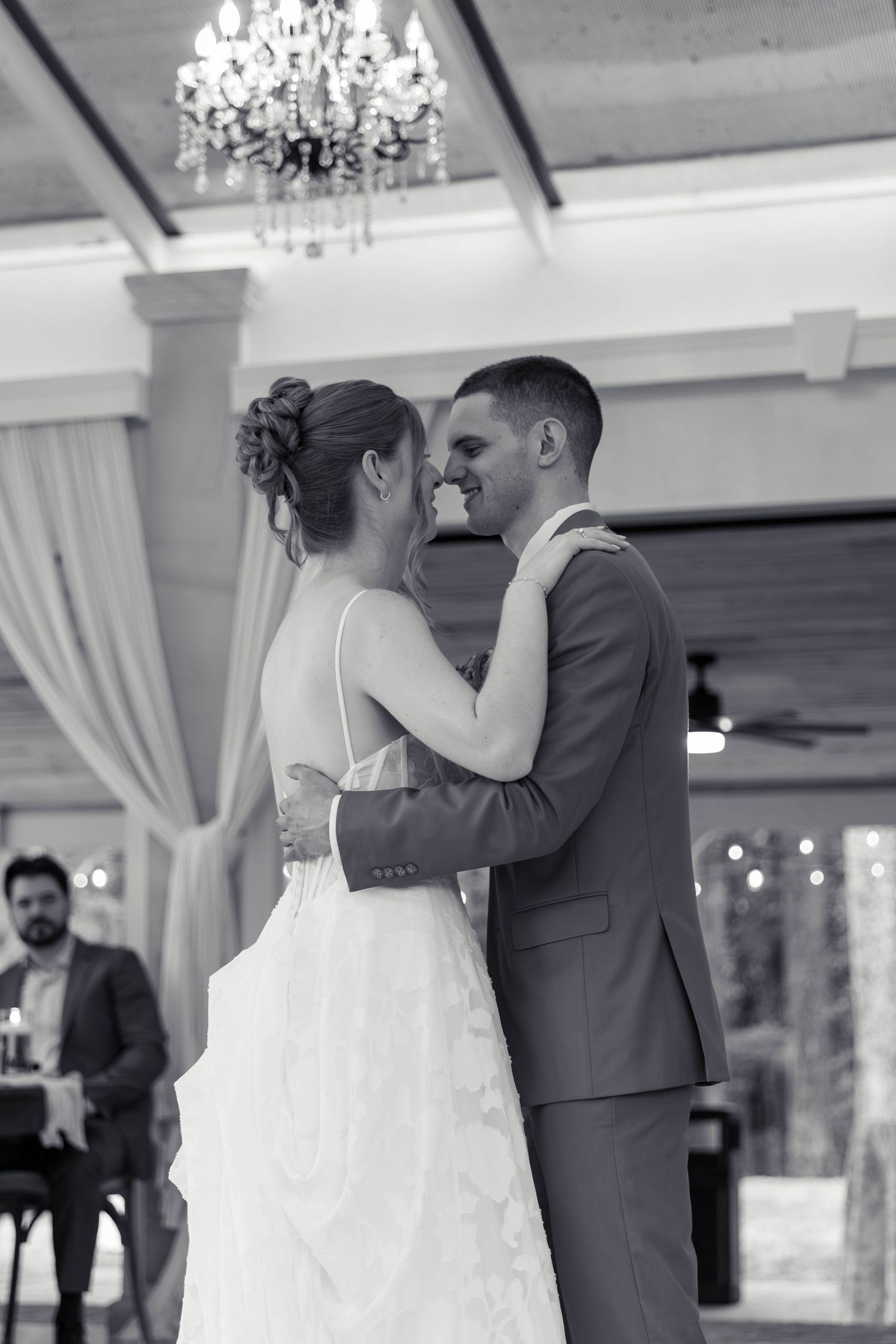 Newlyweds sharing first dance in elegant venue, smiling at each other under chandelier.