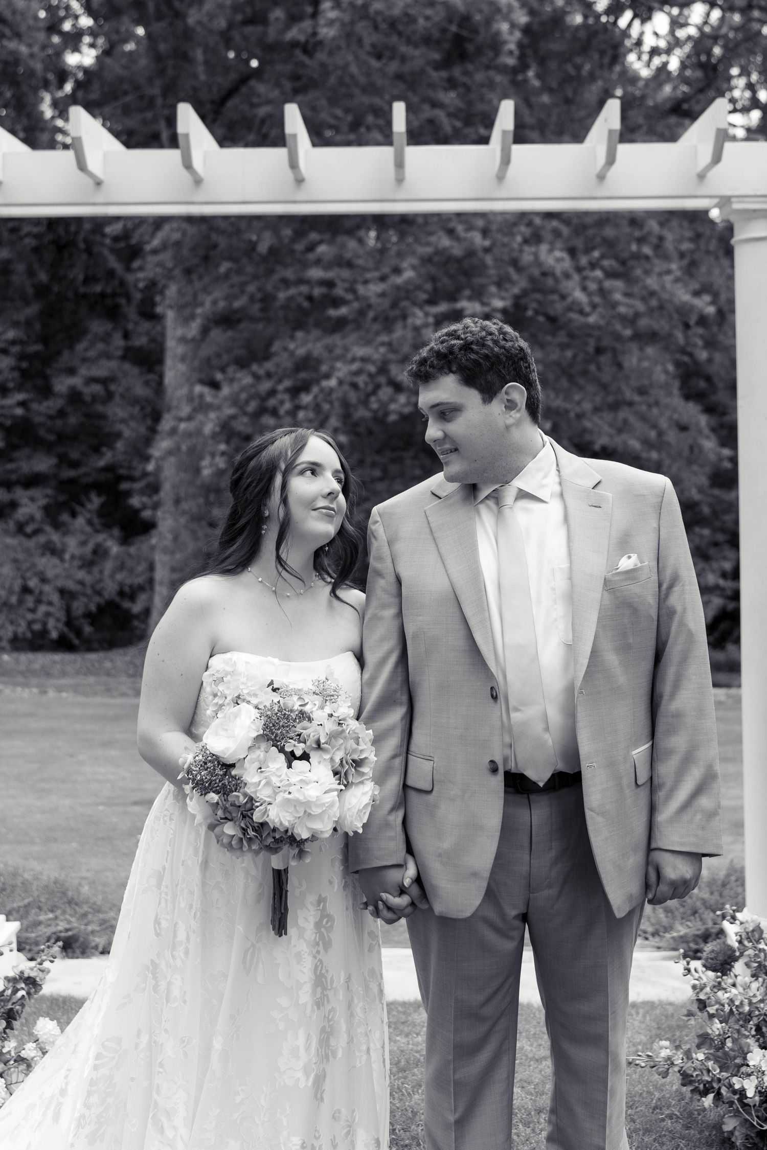 Black and white portrait of bride and groom holding hands under a white pergola outdoors.