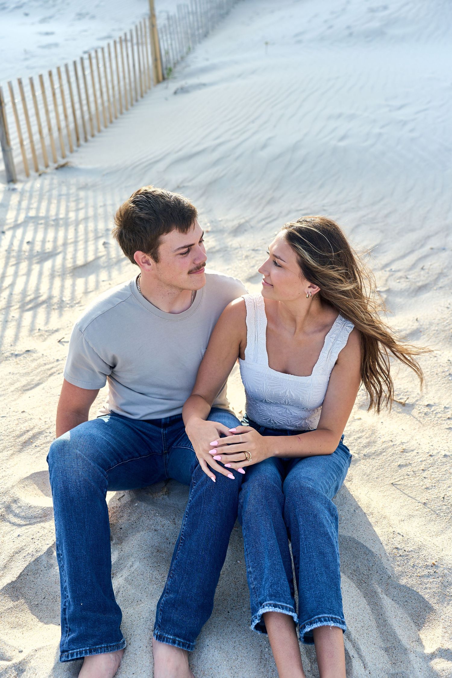 Young couple sitting together on sandy beach with wooden fence in background, sharing intimate moment at sunset.