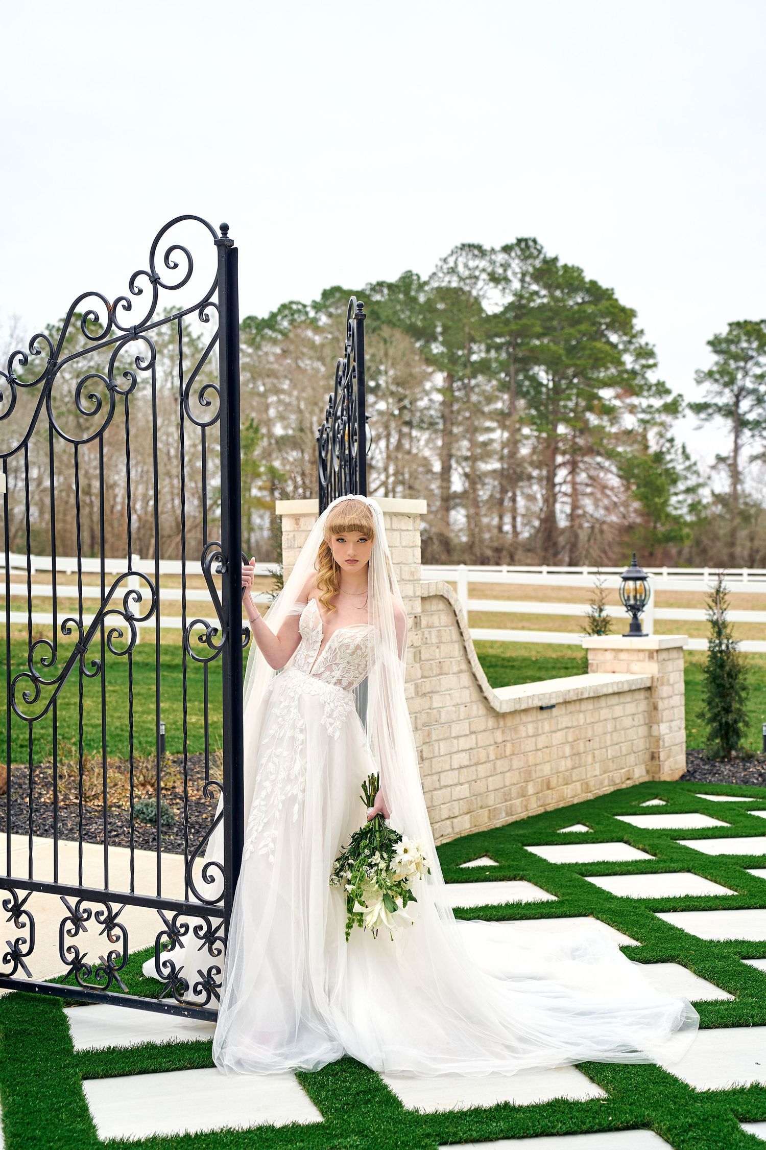 Bride in flowing lace gown stands by ornate wrought iron gate and brick wall on checkerboard turf.