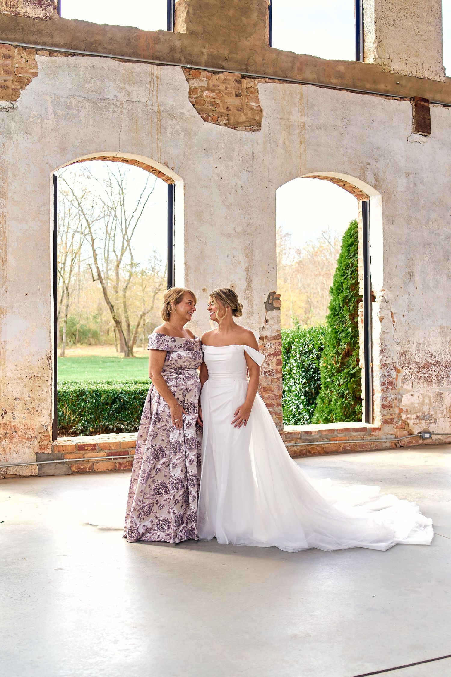 Two people in formal dresses stand together in an elegant stone structure with arched windows overlooking a garden.